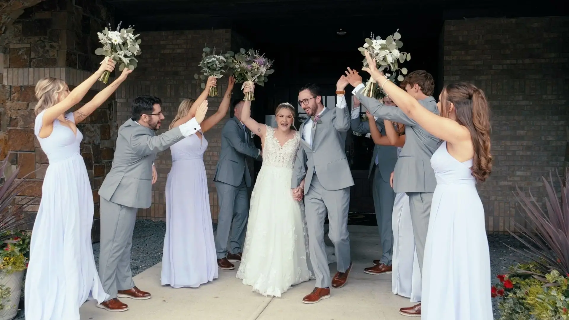 Bride McKenna and groom Jacob walk excitedly under an arch of raised bouquets and joyful hands formed by their bridesmaids and groomsmen at The Union Pavilion at Railside. McKenna wears a flowing white gown, and Jacob is in a light gray suit.