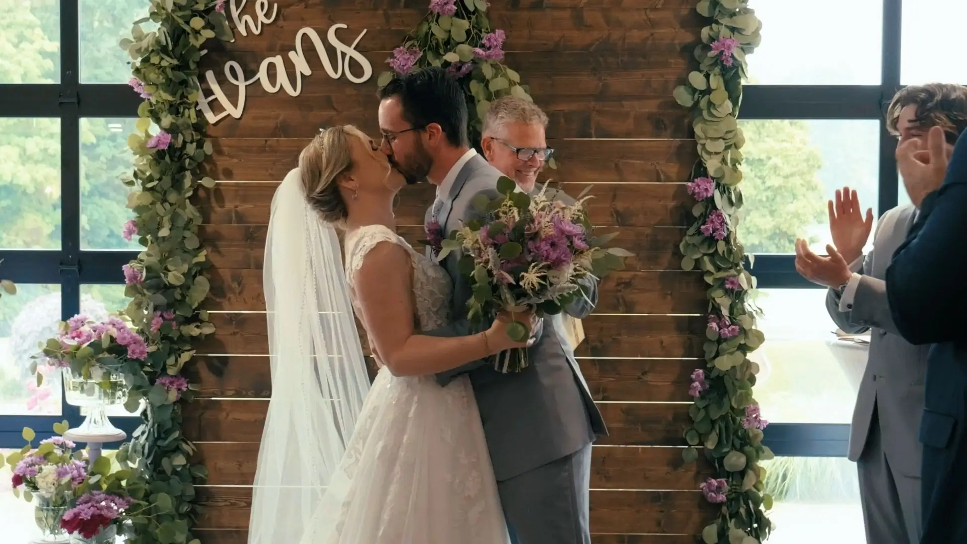 Bride and groom share their first kiss as a married couple in this still from their wedding video at the Union Pavilion at Railside in Byron Center, Michigan. Floral decorations and cheering guests can be seen in the background.