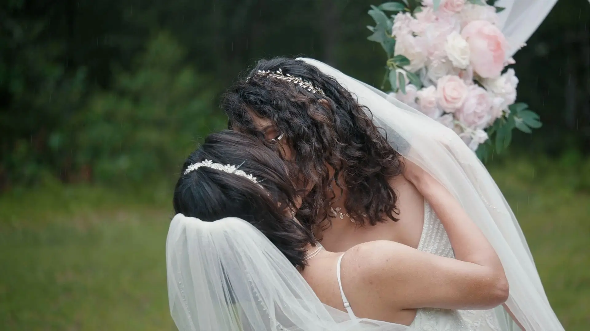 A newlywed couple, Irene and Claudia, share a kiss outdoors during their wedding ceremony at an AirBnB. Both are wearing white wedding dresses with veils and delicate headpieces. A bouquet of pink and white flowers is visible in the background.