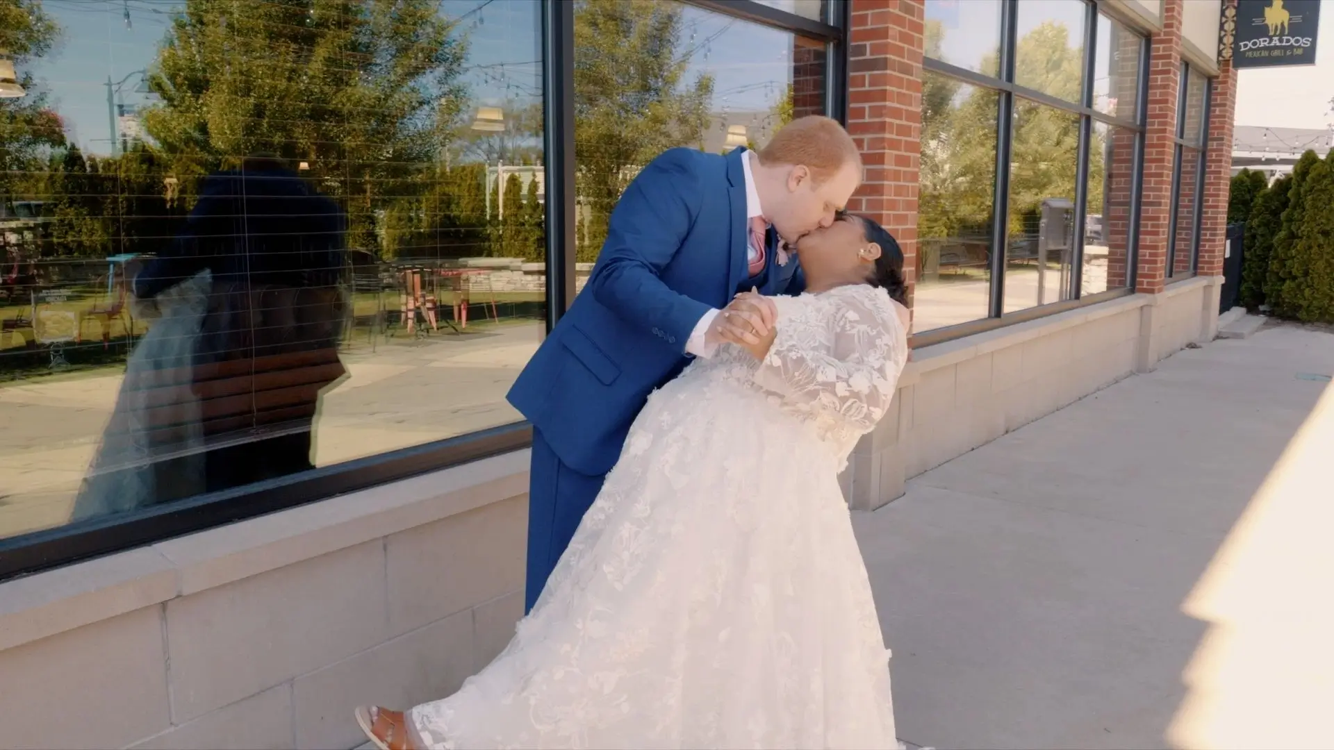 Eveli and David share a kiss and dance outside a building at Terra Square. The groom is wearing a blue suit, and the bride is in a white lace wedding dress. The couple is reflected in the window behind them, and greenery is visible through the window.