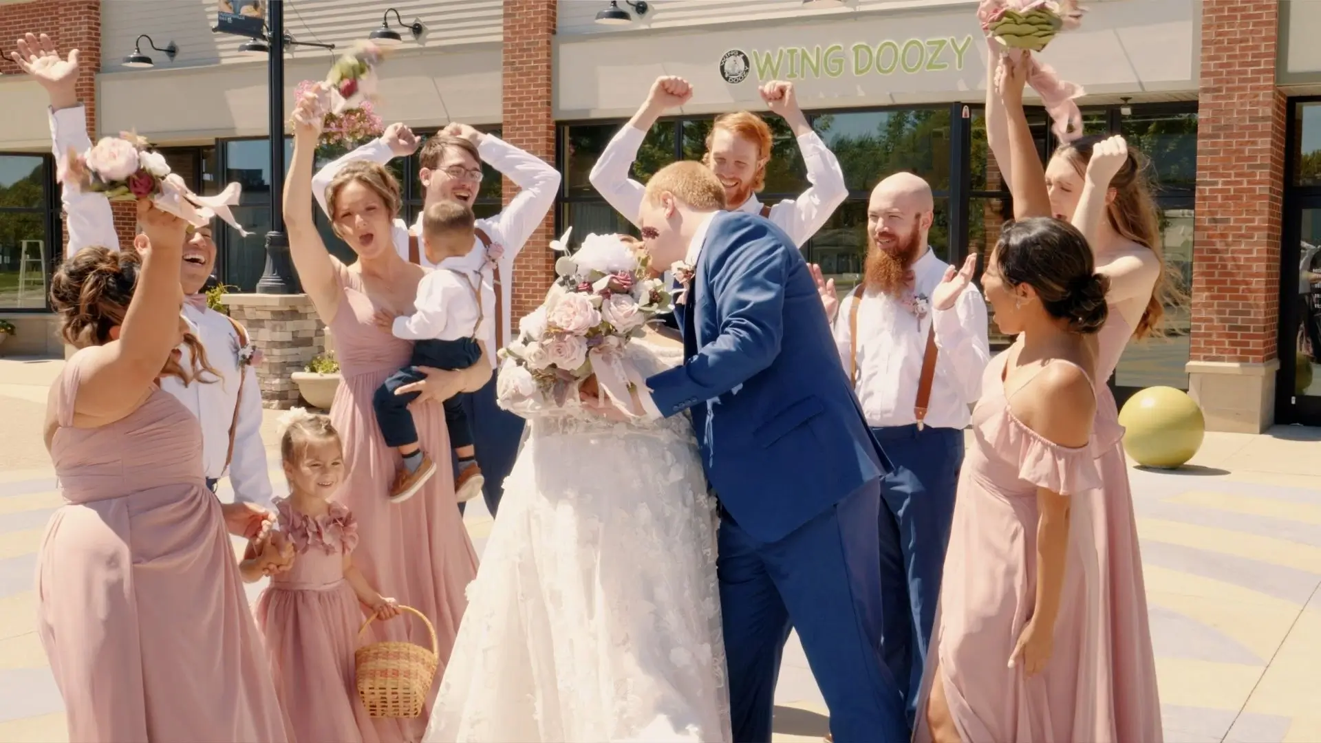 A wedding party celebrates outside Terra Square. The bride, Eveli, in a white dress, and groom, David, in a blue suit, share a kiss while surrounded by their joyful bridesmaids in pink dresses and groomsmen in white shirts and suspenders, holding bouquets.
