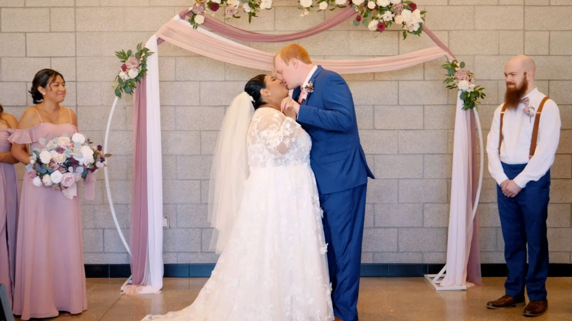 Eveli and David share a kiss during their wedding ceremony. Eveli is wearing a lace wedding gown and veil, and David is in a blue suit. Bridesmaids in lavender dresses and groomsmen wearing suspenders stand on either side under a floral arch.