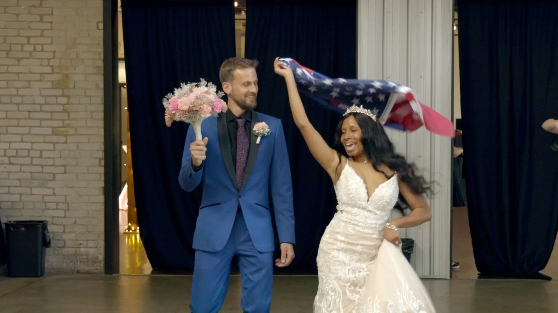 Bride Valérie and groom Nicholas share a joyful moment at their wedding in Studio D2D, Grand Rapids. Valérie swirls a flag representing their nationalities, while Nicholas holds a bouquet. The couple's happiness is evident as they celebrate their union.