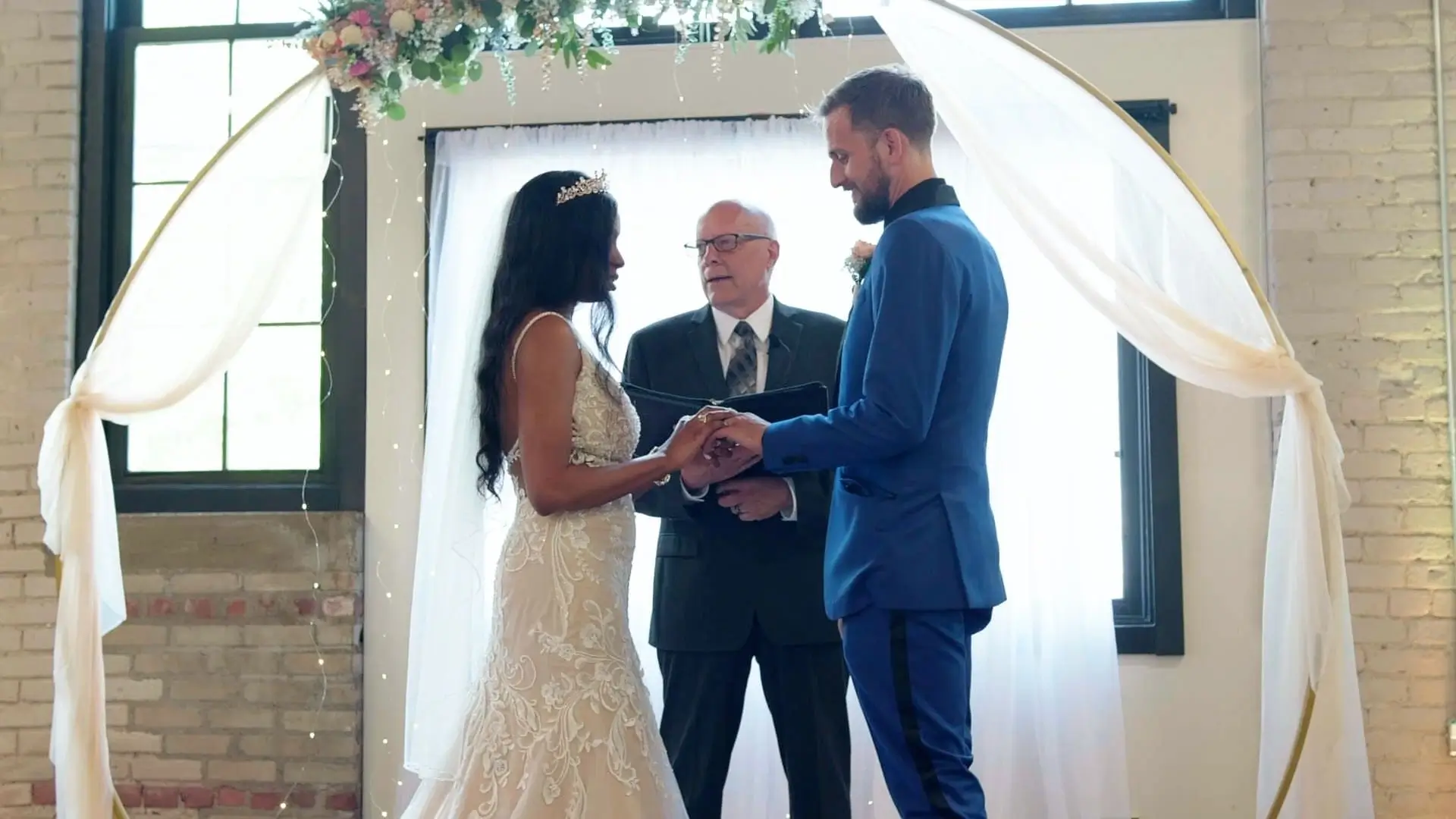 Valérie and Nicholas exchange vows during their wedding ceremony at Studio D2D in Grand Rapids on May 18, 2024. The bride wears a white lace gown, while the groom wears a blue vegan suit. The officiant stands between them under a floral arch.