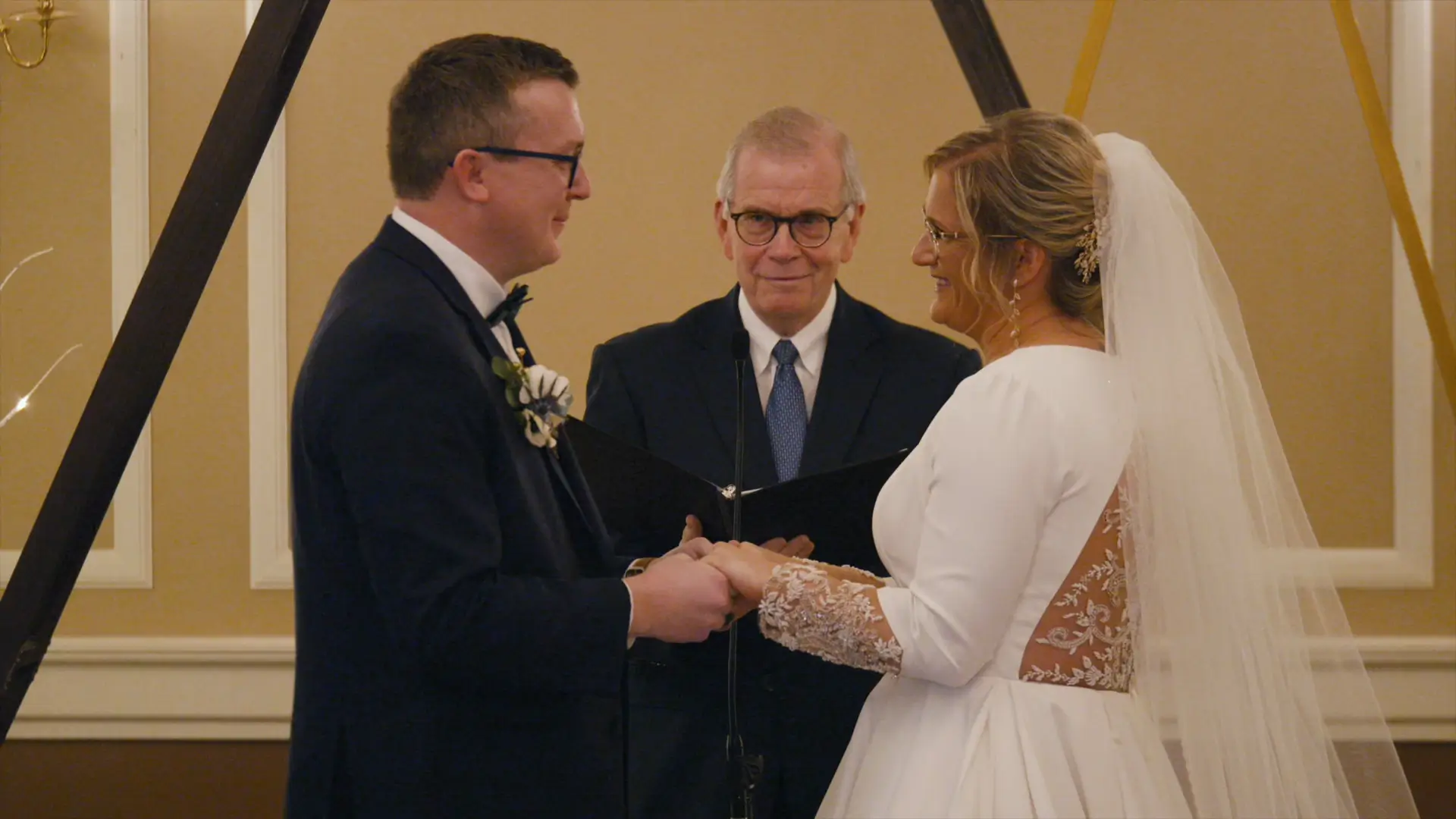 Bride and groom exchanging vows at their wedding in Jackson, Michigan, with an officiant standing between them. The couple is smiling and holding hands in an intimate ceremony setting.