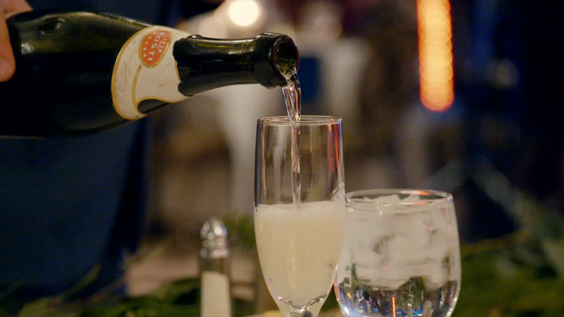 Champagne being poured into a flute glass next to a glass of ice water, set against a blurry, warmly lit background.