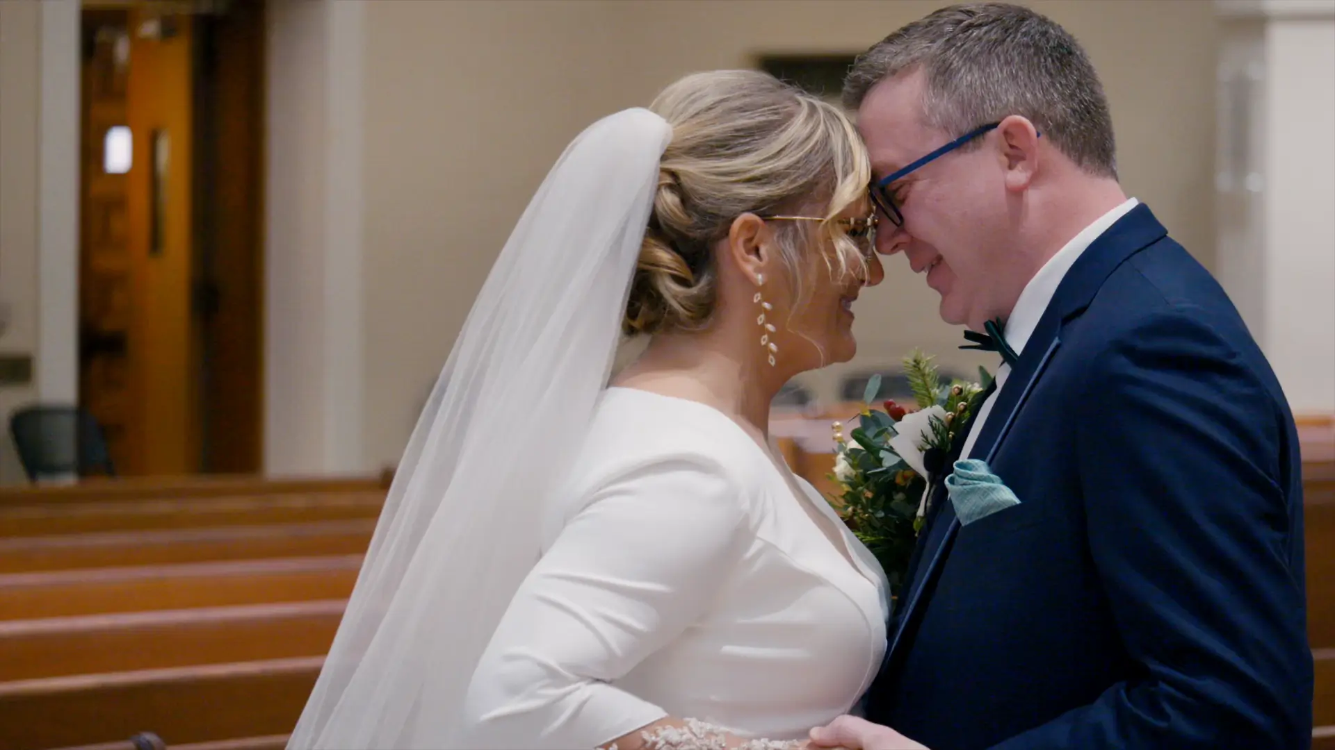 Bride Billie and groom Stephen share a tender moment in Queen of the Miraculous Medal Church in Jackson, Michigan. She wears a white gown with a veil, while he’s in a navy tuxedo with teal accents, holding her bouquet.