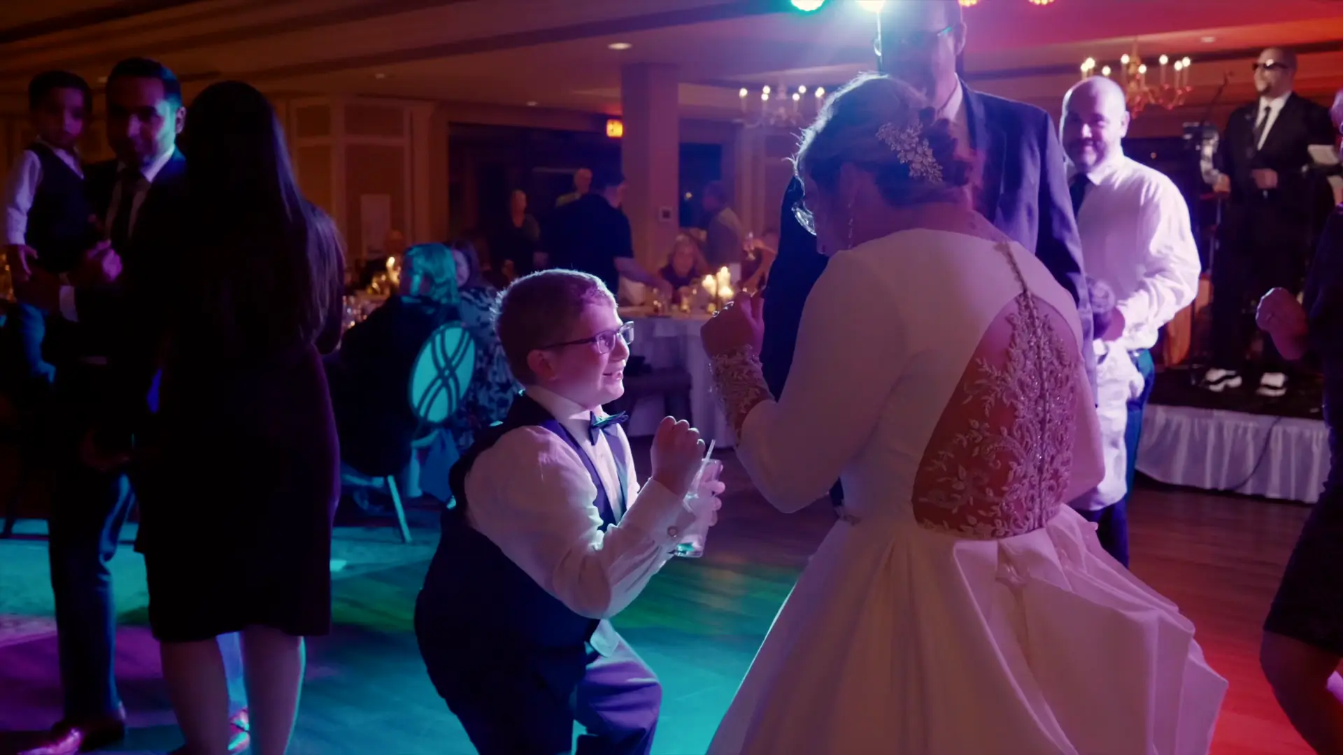 Bride and young boy share a joyful dance at a lively Michigan wedding reception, warmly lit with colorful lights, surrounded by guests and musicians in the background.