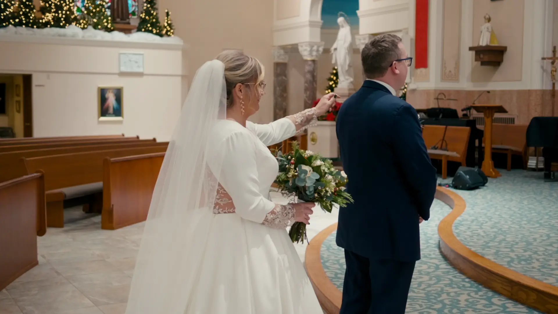 Bride Billie taps groom Stephen's shoulder during a first look at their wedding in Queen of the Miraculous Medal Church, Jackson, Michigan. The church interior is adorned with festive greenery and lights, capturing a heartfelt Michigan moment.