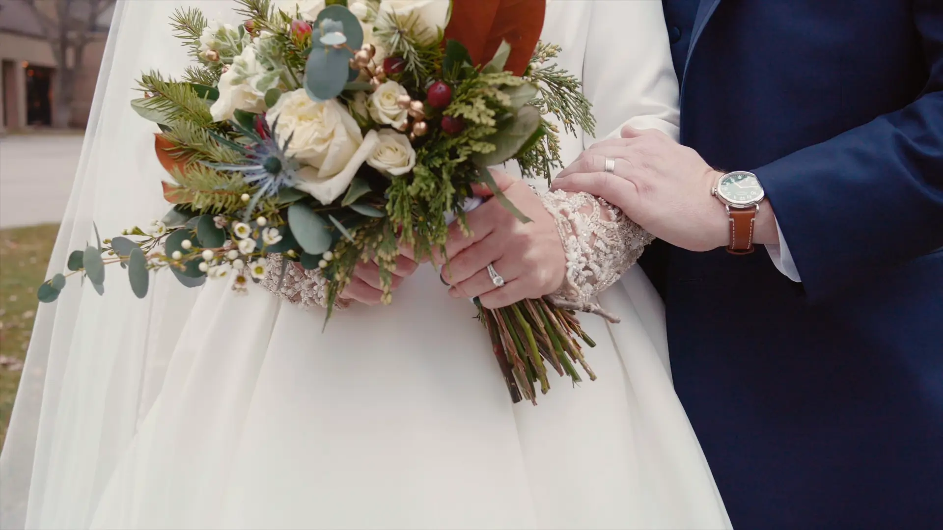 In this snapshot from Billie's and Stephen's wedding video, the couple is seen holding a vibrant bouquet. The bride's lace sleeve is visible, and their hands, both adorned with rings, are gently intertwined, capturing a moment of love in Jackson, Michigan.