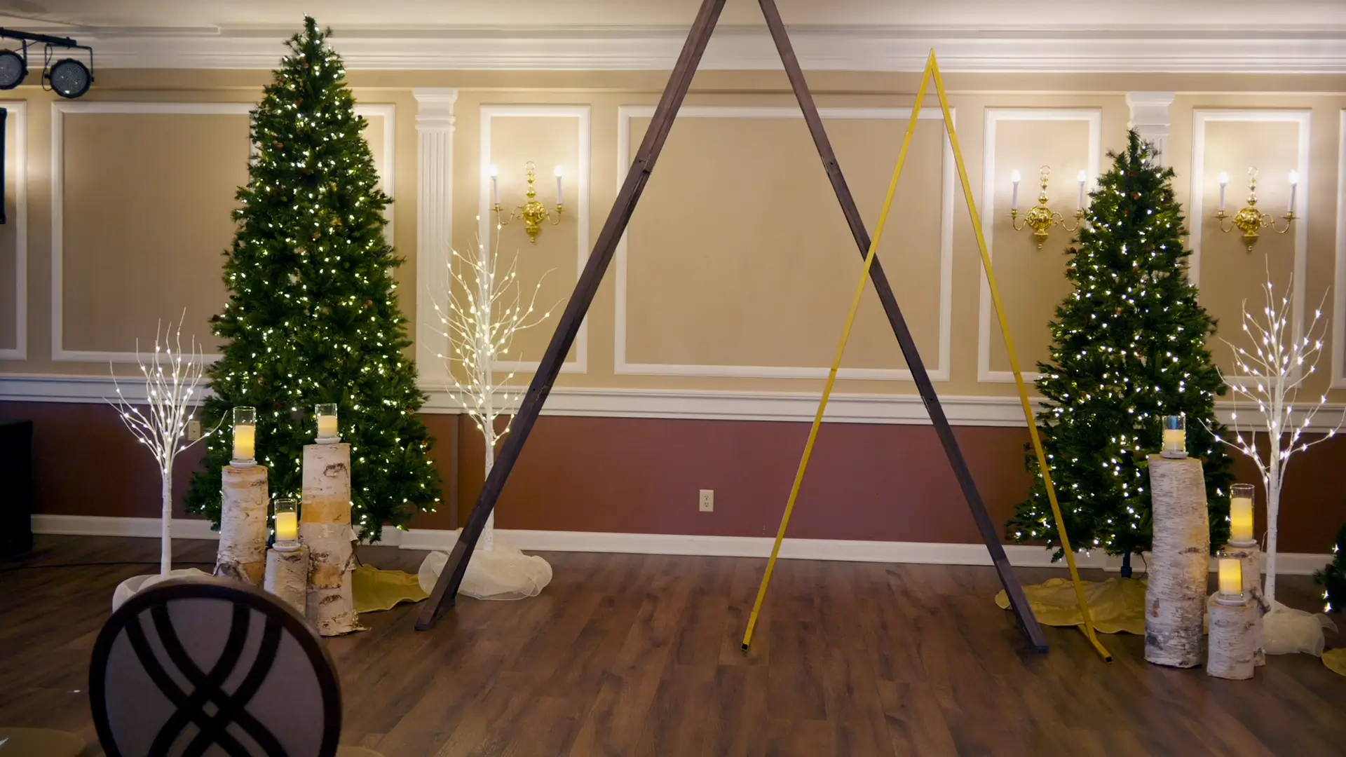A decorated wedding venue in Jackson, Michigan, featuring lit Christmas trees, birch log candle holders, and a wooden arch in a warmly lit room with elegant wall sconces.
