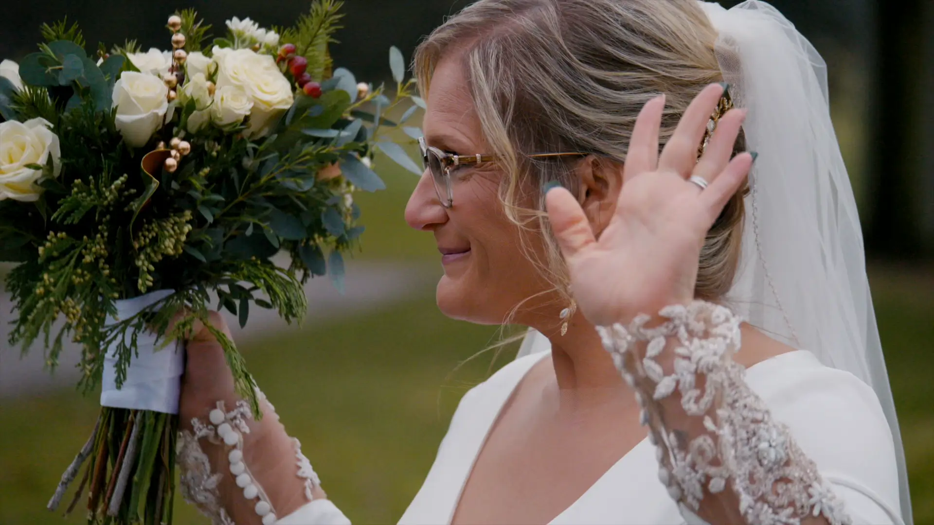 In this still from their wedding videography, Billie, holding a bouquet of white roses and greenery, smiles with a gentle wave. The intricate lace on her sleeves complements her elegant glasses and veil.