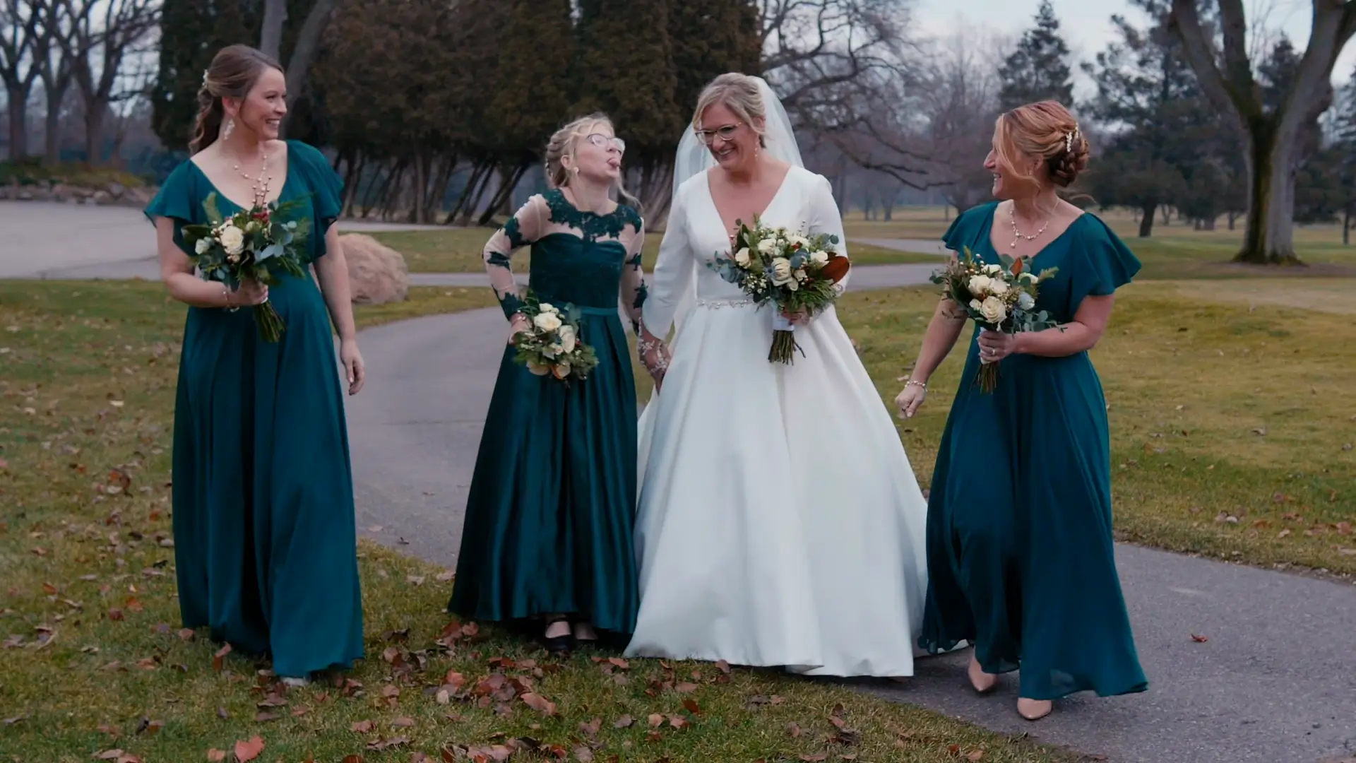 Bride Billie in a white gown and bridesmaids in teal dresses share a joyful moment on a walkway, holding bouquets. This candid scene captured by the wedding videographer highlights laughter and togetherness at Billie and Stephen's wedding in Jackson.