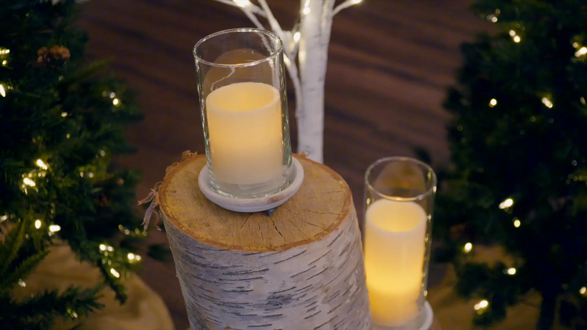 Warm lighting from candles in glass holders on birch logs, surrounded by softly lit evergreen trees, creates a cozy ambiance. Perfect for a romantic Michigan wedding setting.