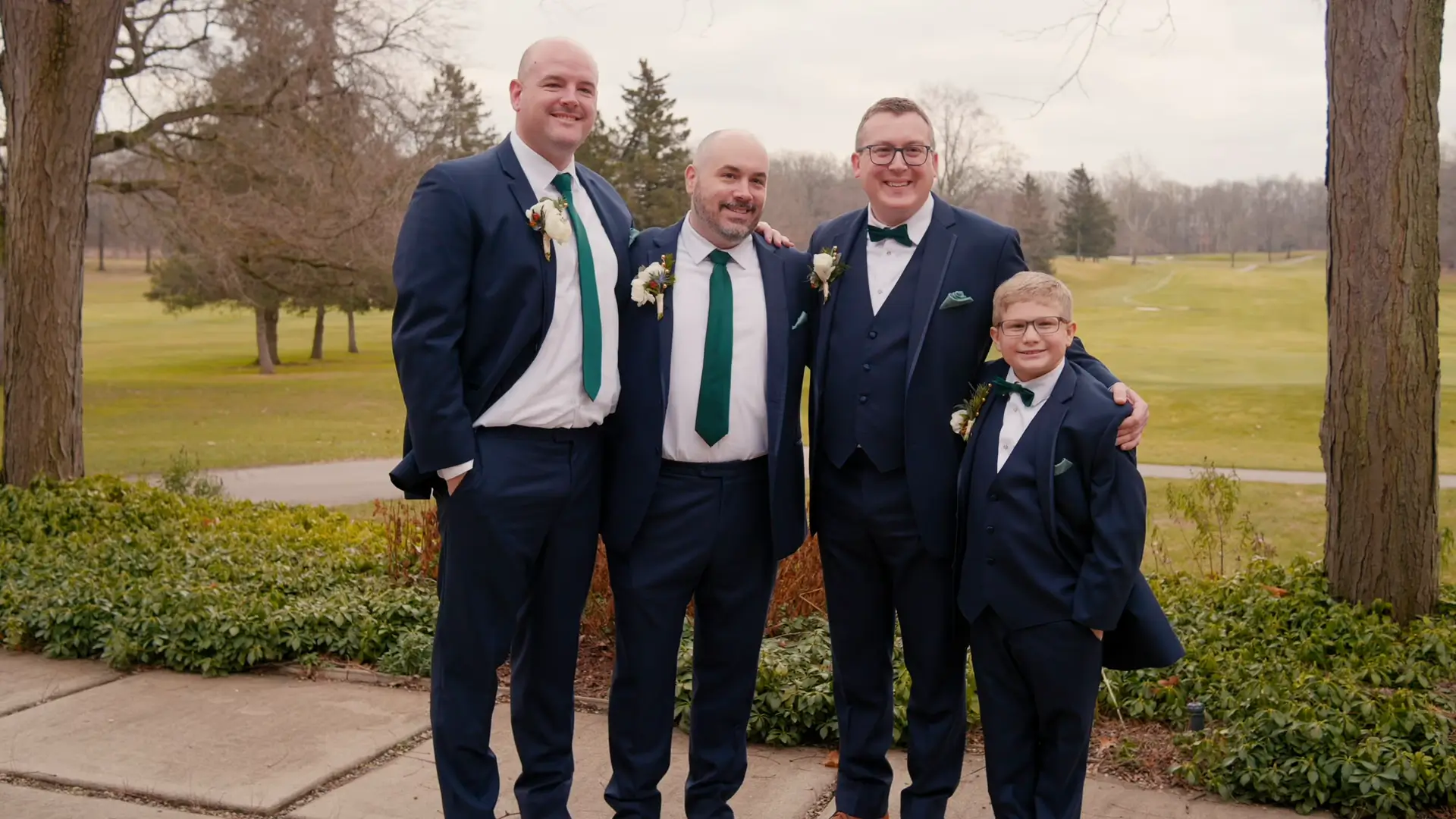 Groomsmen and a junior groomsman in matching suits smile together in front of a lush golf course in Jackson, Michigan, capturing a joyful wedding moment.