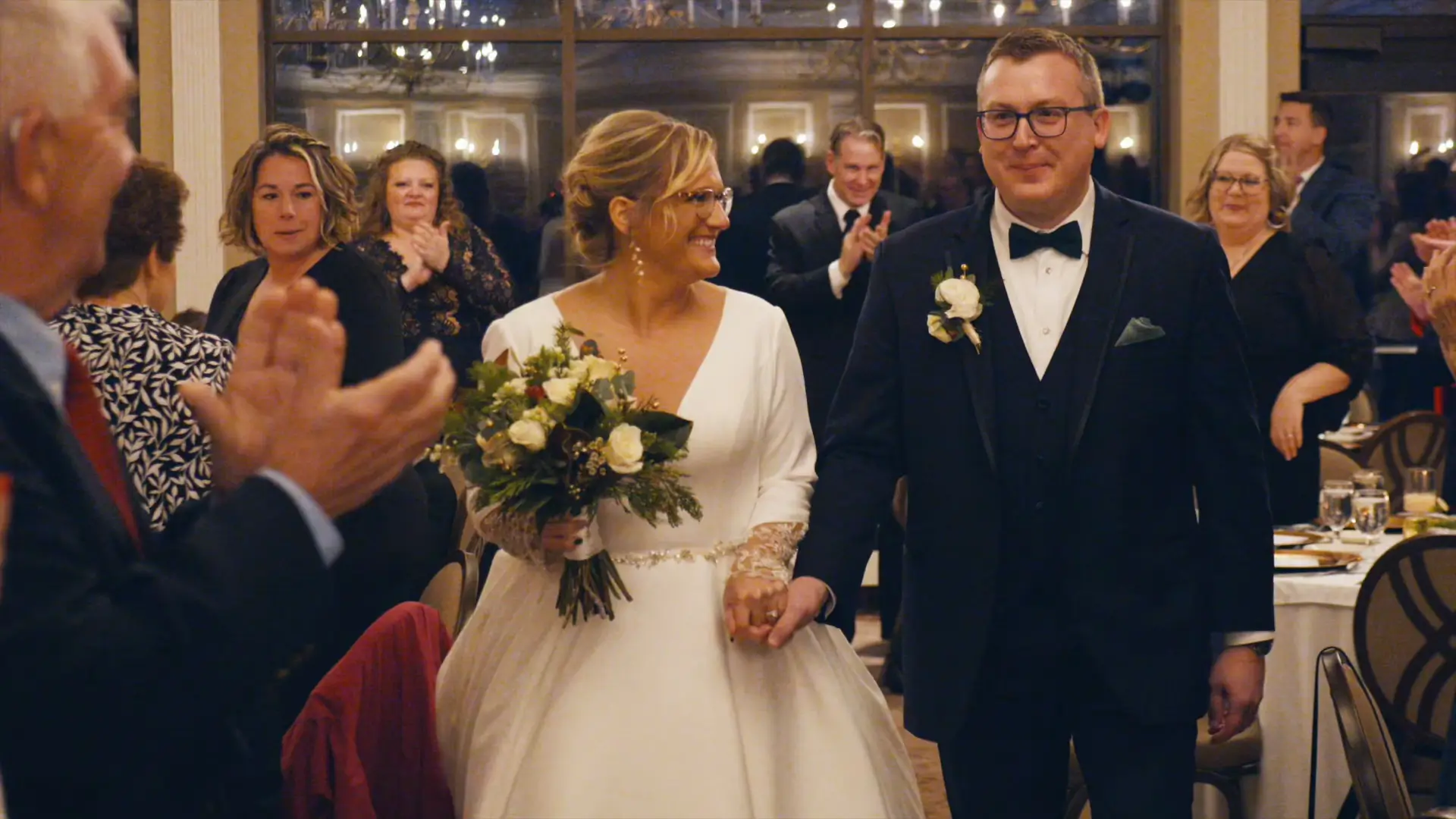 Billie and Stephen walk hand-in-hand through a crowd clapping and smiling at their wedding reception in Jackson, Michigan. Billie, the bride, is in a white dress and holds a bouquet; Stephen, the groom, wears a black suit with a bow tie.