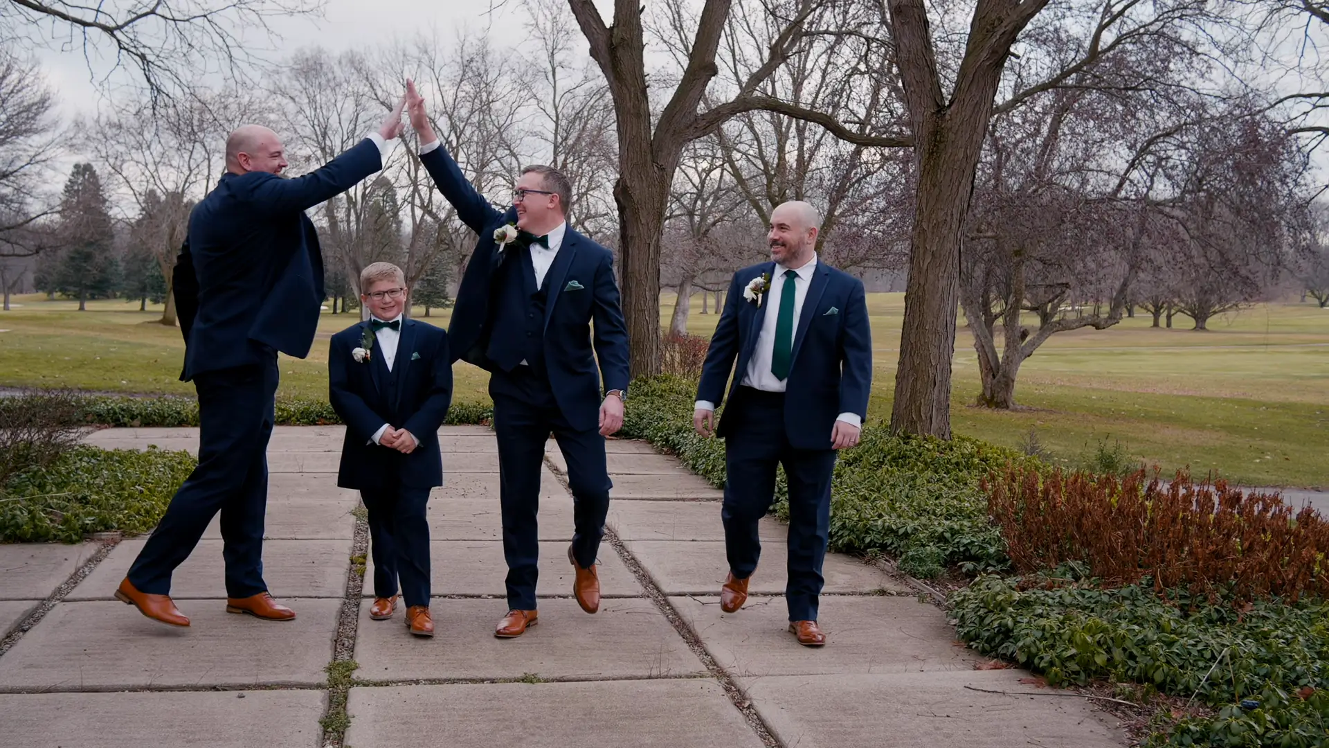 Groomsmen in navy suits high-fiving joyfully, with a young boy in a matching suit smiling nearby. Leafless trees and a wintry Michigan landscape create a serene background.