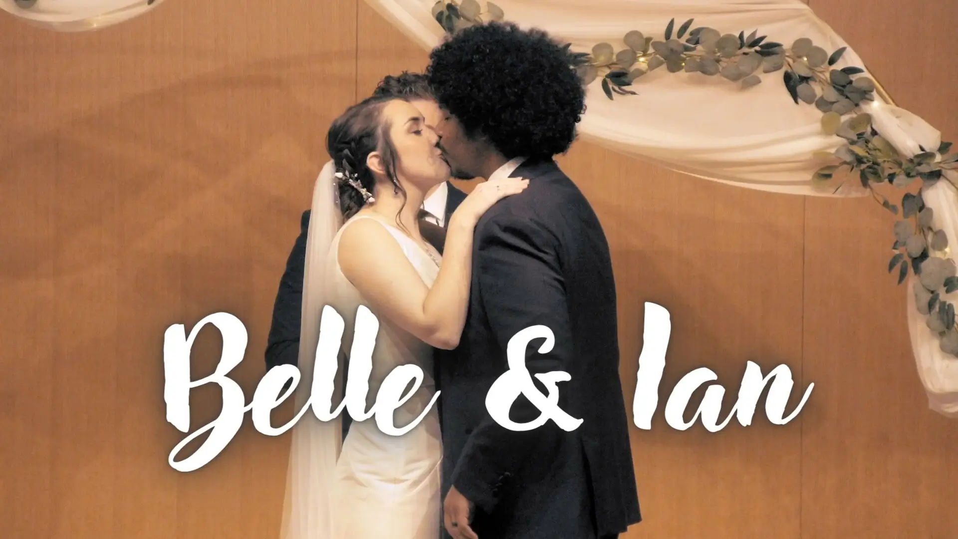Belle and Ian share a kiss in front of a floral-draped arch during their wedding ceremony at the Grand Rapids Art Museum in Michigan. Large white text at the bottom reads "Belle & Ian."