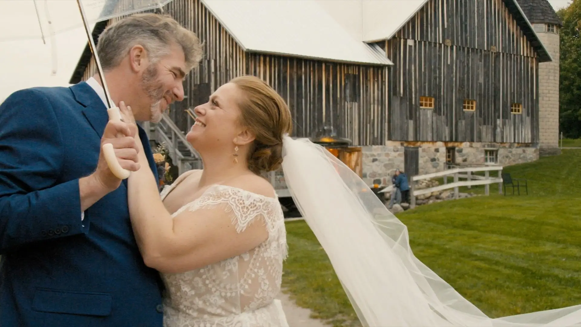 Beth and Aaron, the bride and groom, smile while dancing under an umbrella in front of the rustic Starry Night Barn & Studios in Suttons Bay, Michigan. The bride's veil flows in the wind, and the groom wears a dark blue suit.