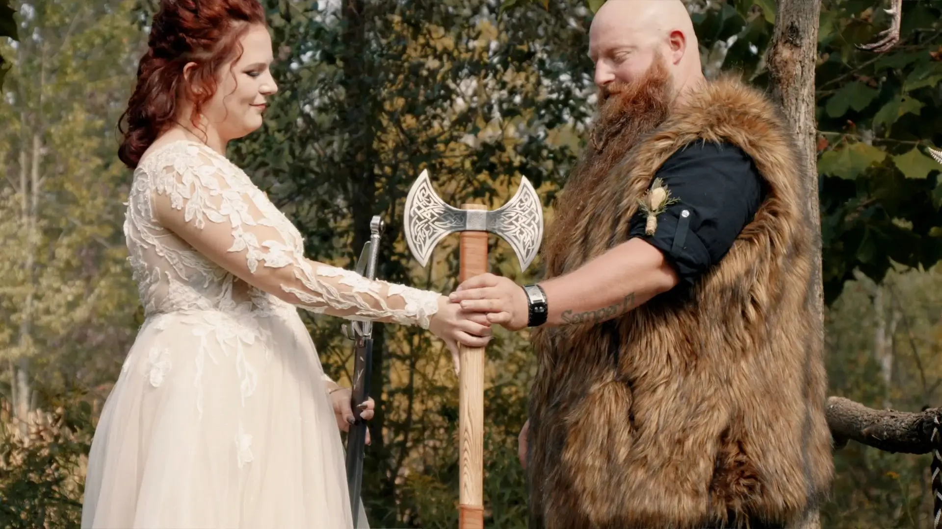 Bride Milah and groom Keith perform a hand fasting ritual during their Viking-themed backyard wedding. Milah wears a lace gown and Keith has a fur vest. They hold a double-headed axe, symbolizing their union, with a green forest in the background.