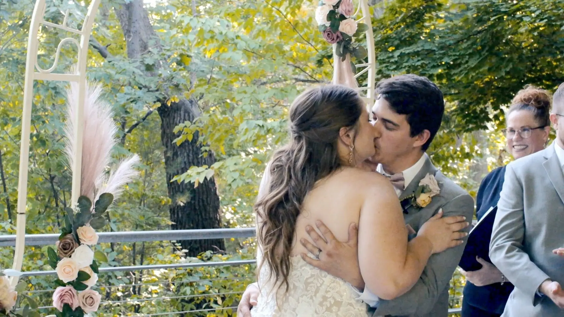 Bride and groom share a kiss during their wedding ceremony amid lush greenery at The Bissell Tree House in Grand Rapids, Michigan. The bride wears a strapless gown, and the groom is in a light gray suit while the officiant and wedding party smile.
