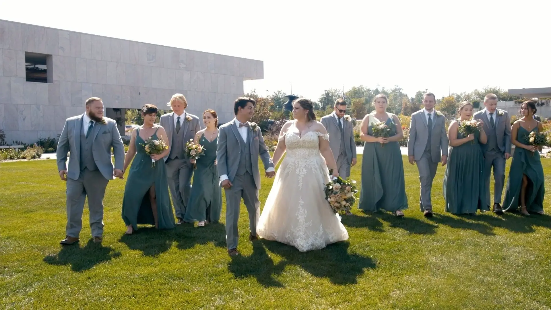 Bride and groom, Abigail and Jordan, walk hand-in-hand with their wedding party, dressed in grey suits and teal dresses, across a grassy lawn on a sunny day at Frederik Meijer Gardens in Grand Rapids. The venue's modern building is in the background.