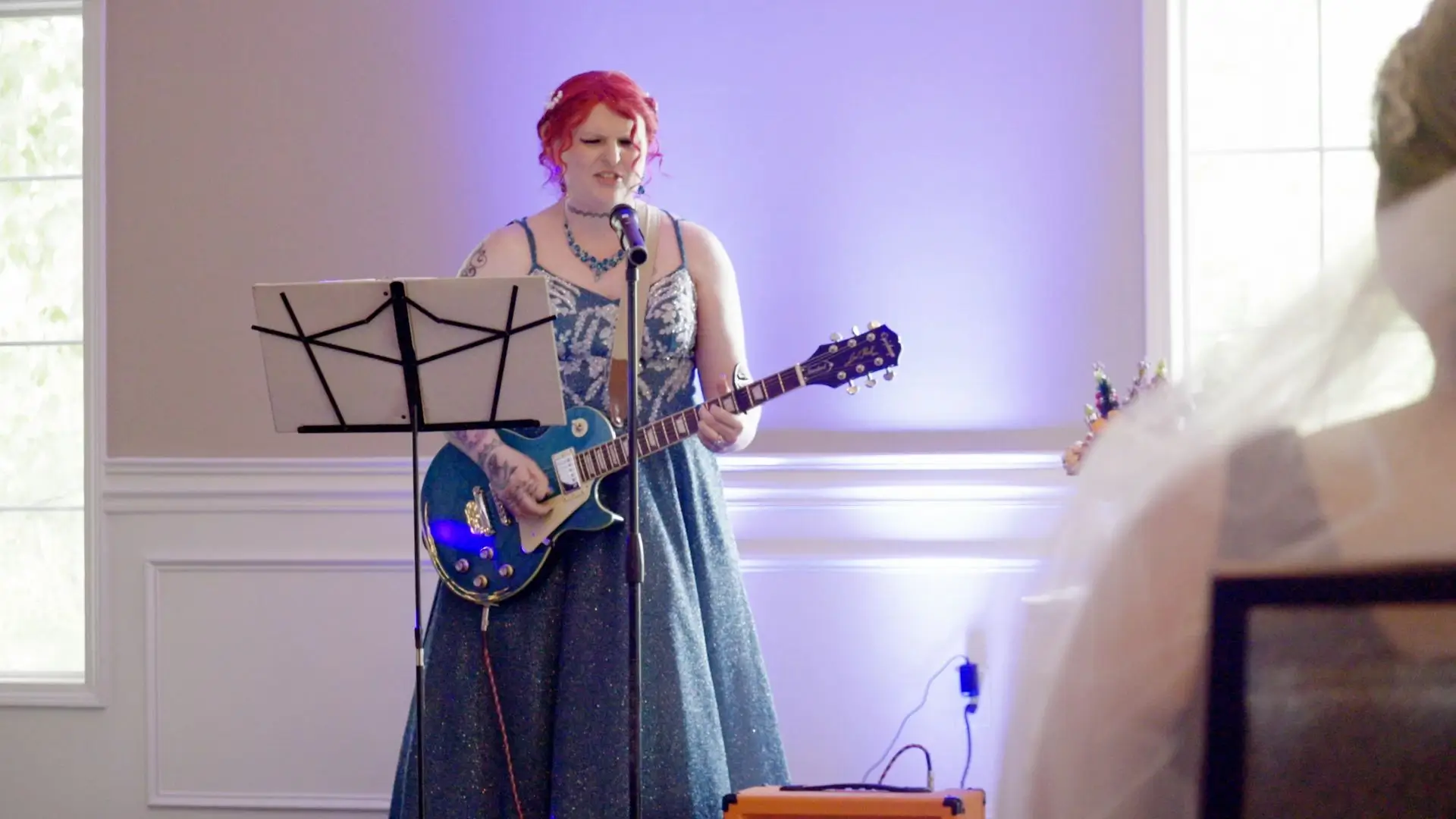 Danni, with red hair, stands on stage singing into a microphone and playing an electric guitar at her wedding to Melissa at The English Inn. She wears a glitter-covered dress and is accompanied by a music stand with sheet music.