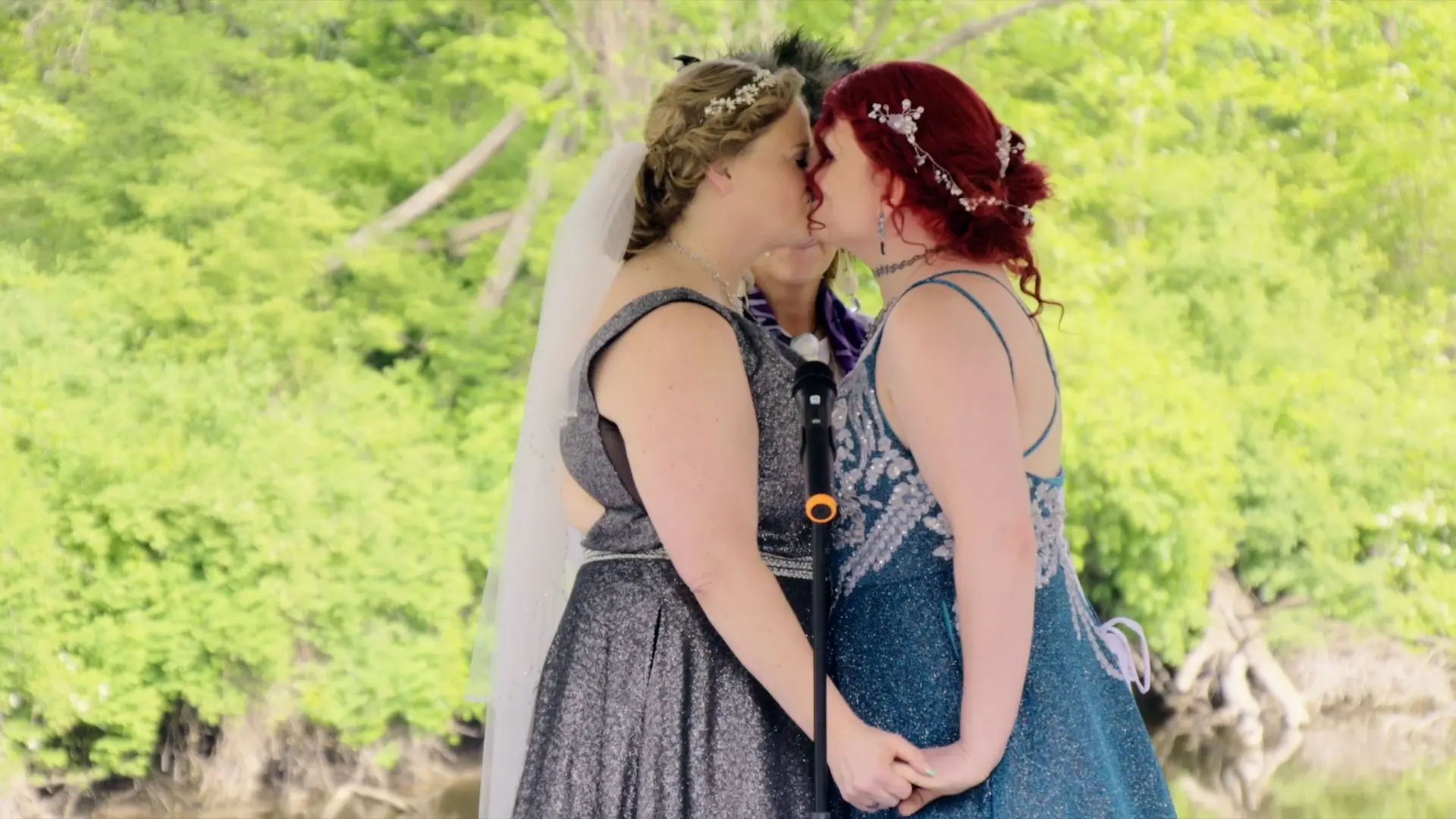 In this still from their wedding video, Melissa and Danni share a kiss at their ceremony in The English Inn. Both brides are in glitter dresses, holding hands with a lush backdrop. The officiant stands in the background.