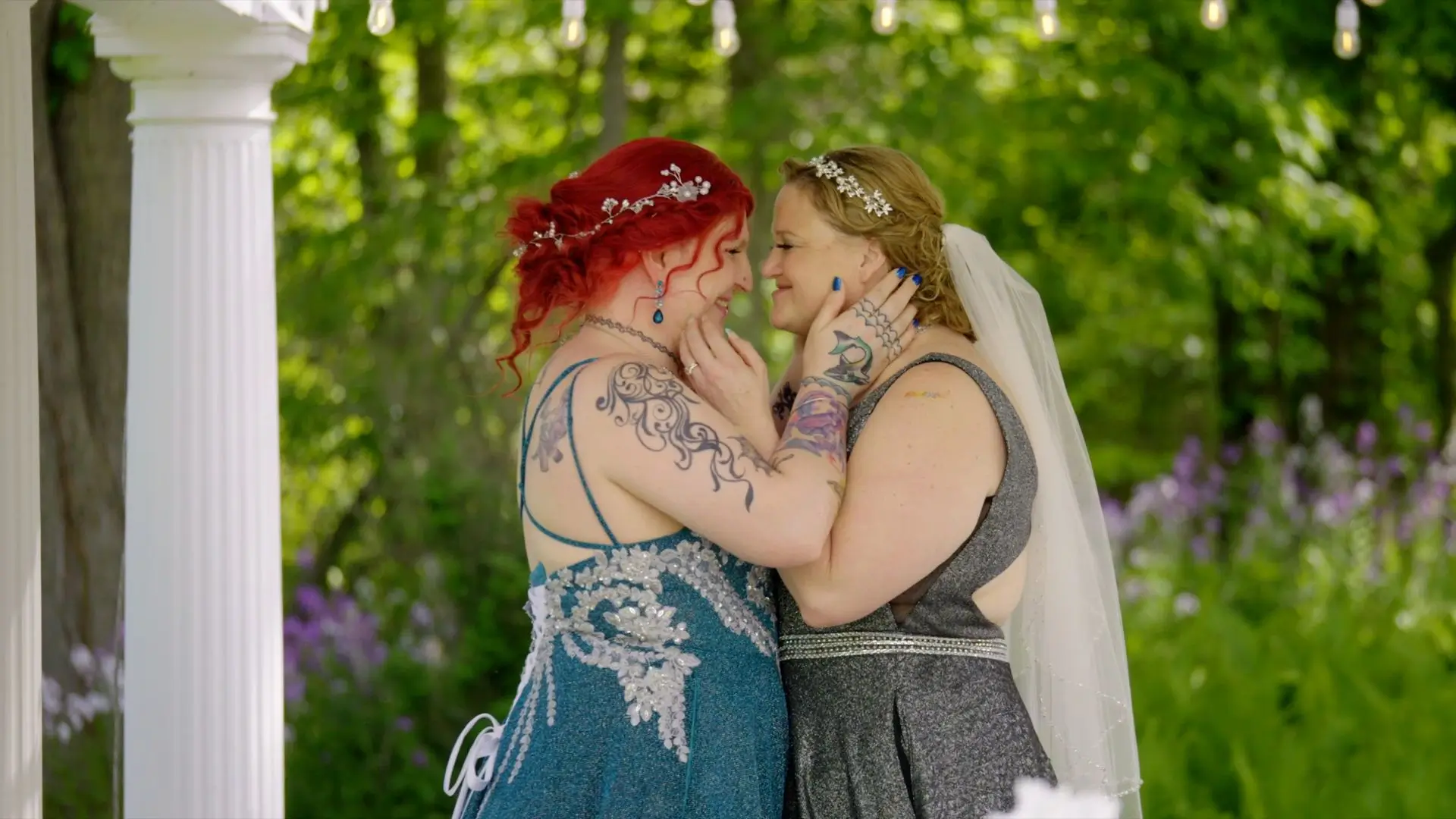 Melissa and Danni, adorned with tattoos and headpieces, share a tender moment during their wedding at The English Inn. Melissa, in a grey dress and veil, and Danni, in a blue dress with red hair, lovingly touch each other's faces.