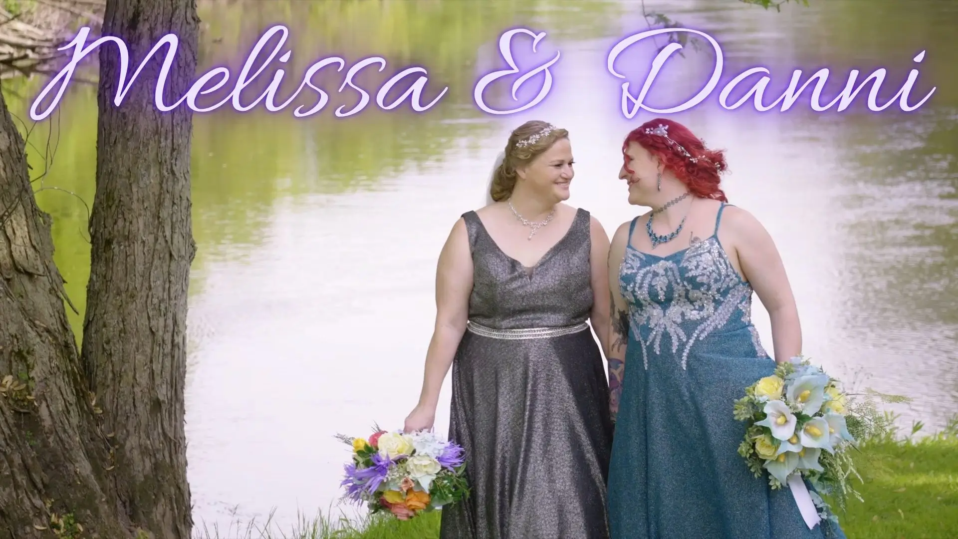Melissa and Danni, dressed in glittering wedding gowns, smile at each other while standing next to a serene lake at The English Inn in Eaton Rapids, Michigan. Both brides hold vibrant bouquets, reflecting joy and the peaceful greenery of Michigan.