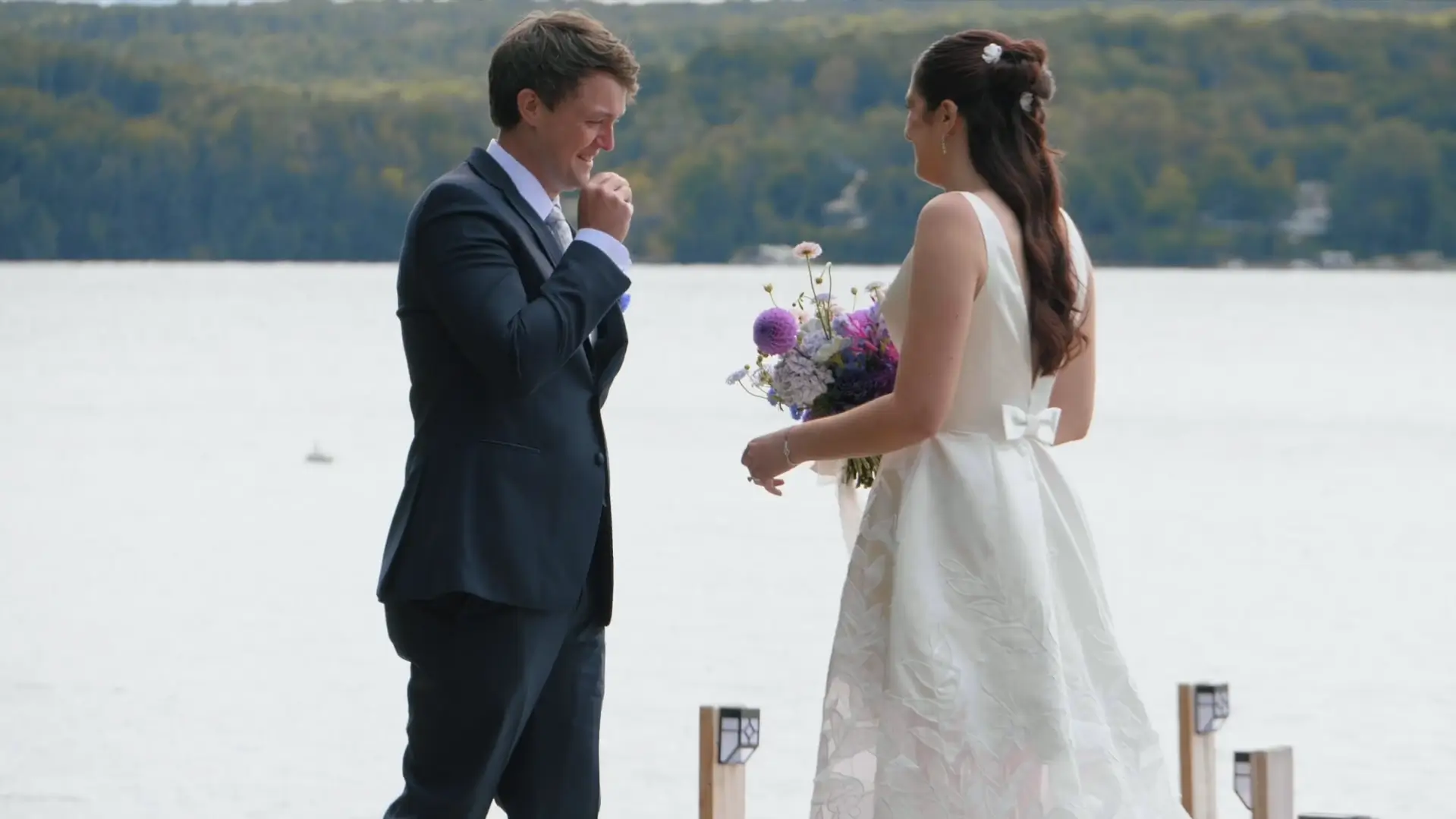 Groom Joe becomes emotional when he sees his bride Chloe during their first look on a dock at Walloon Lake. He wears a blue suit and Chloe wears her wedding dress, holding a vibrant bouquet.
