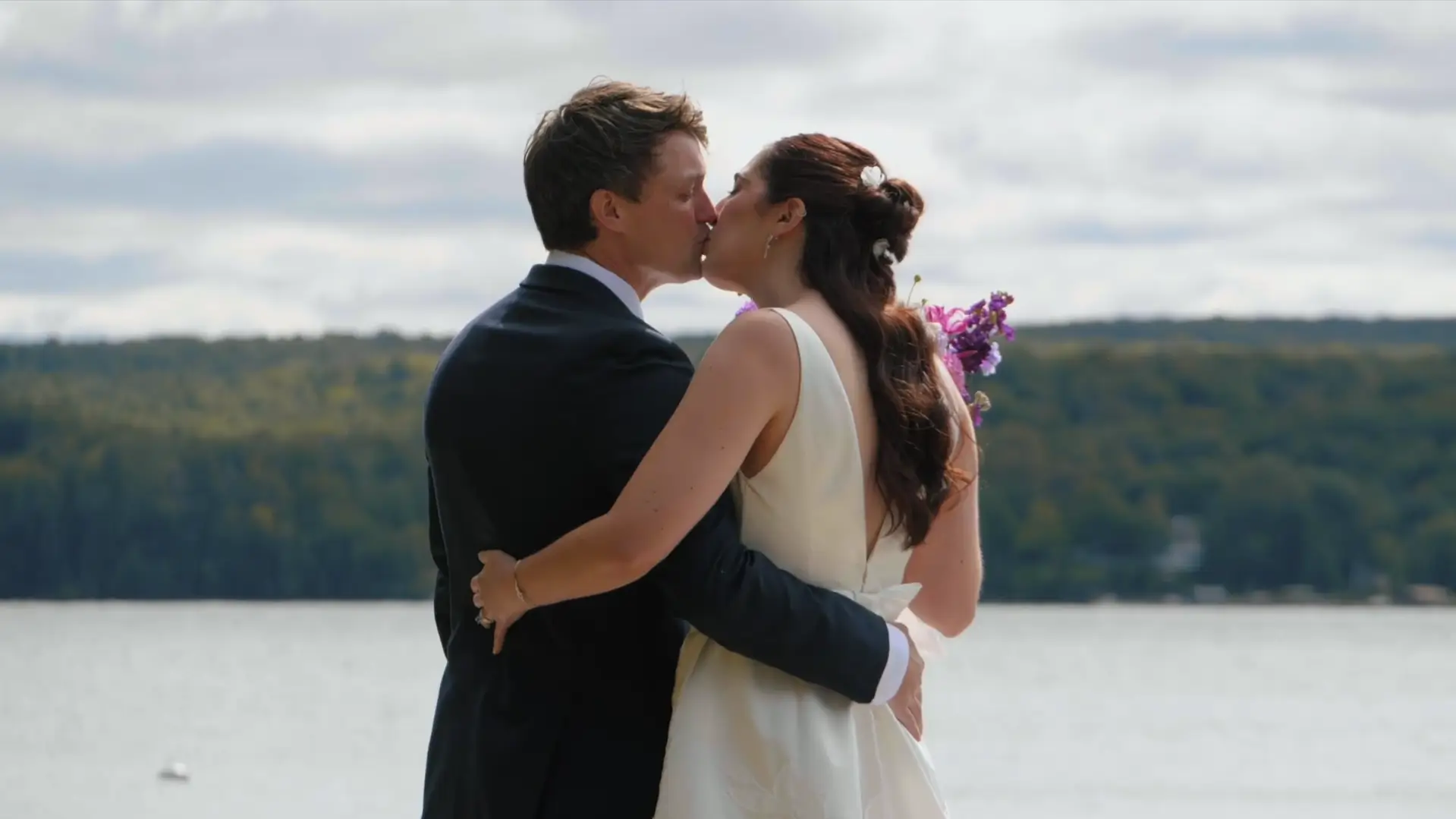 Bride and groom share a kiss by the beautiful Michigan waterfront at Walloon Lake, with lush greenery in the background. The bride holds a vibrant bouquet, and both are elegantly dressed, capturing a romantic moment during their wedding celebration.