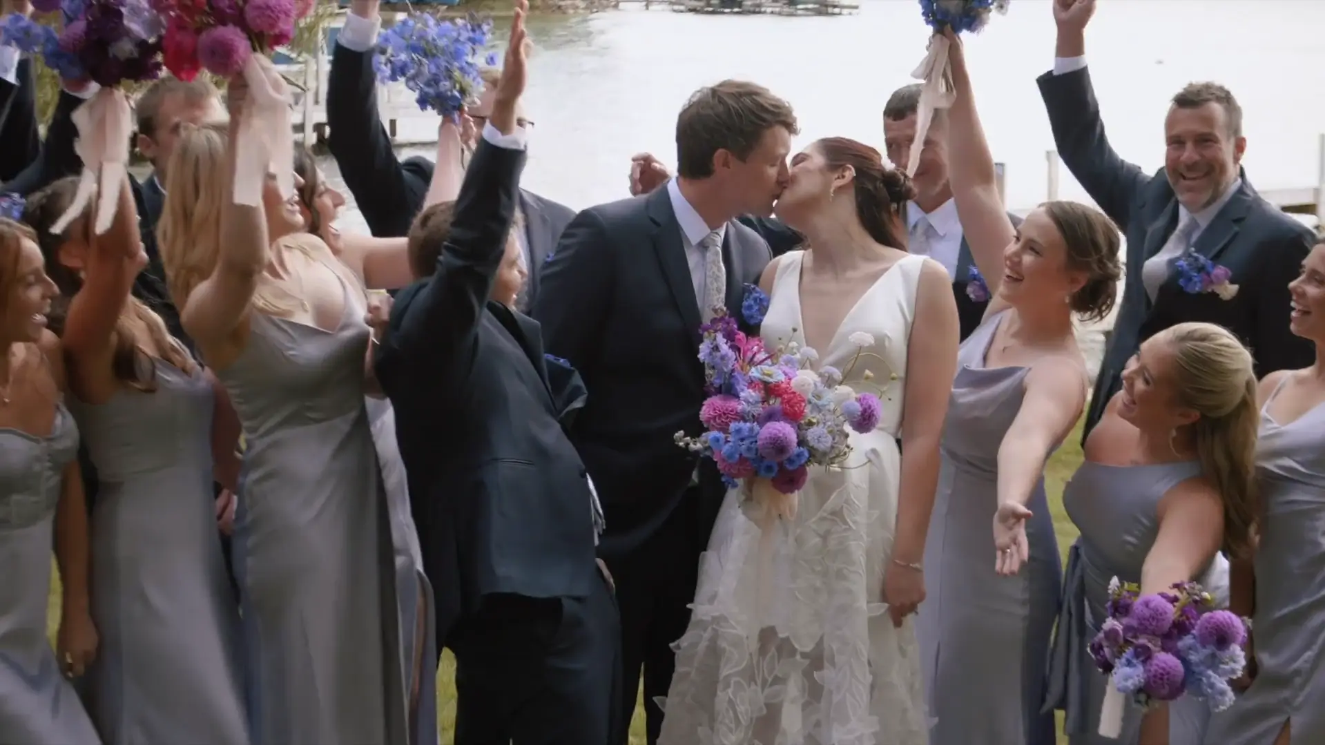 Chloe and Joe share a kiss surrounded by their wedding party, with bridesmaids in lavender dresses and groomsmen in suits, cheering and raising bouquets.