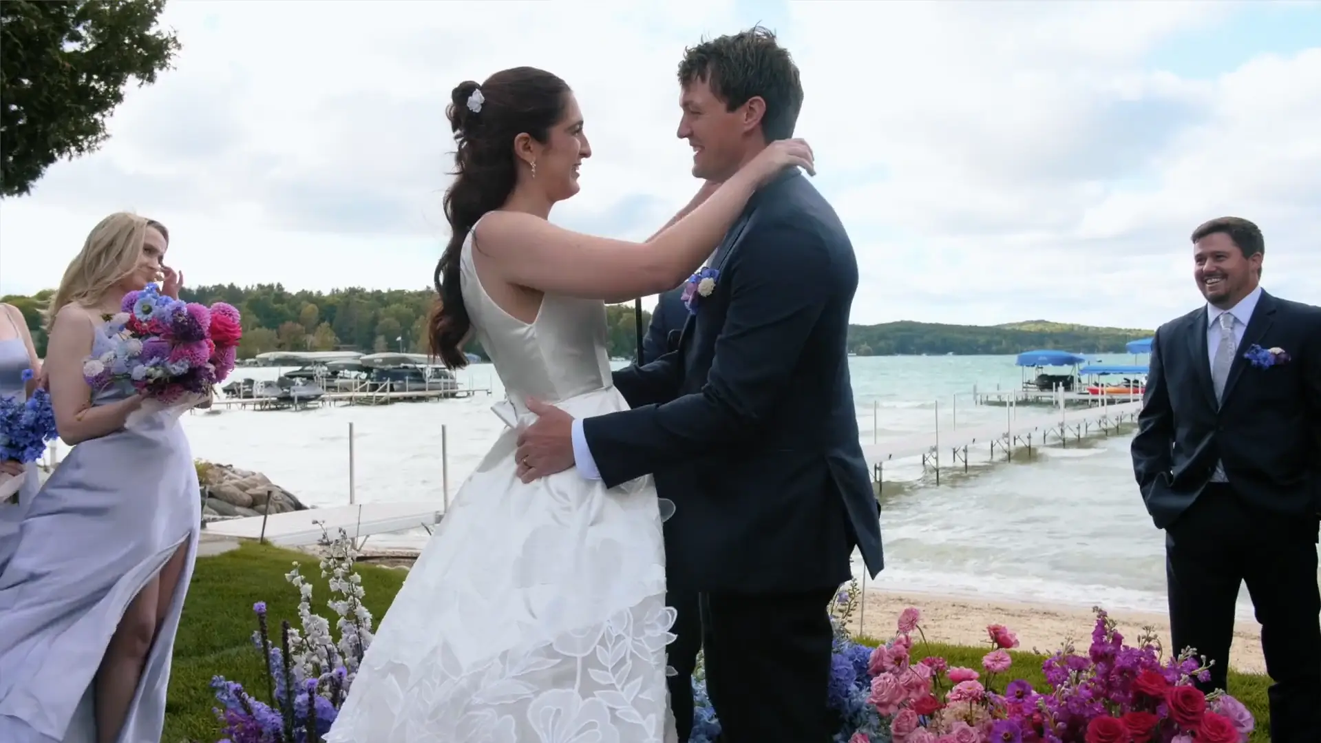 Bride and groom embrace lakeside in Petoskey, Michigan. Bridesmaids in lavender dresses hold vibrant bouquets. A serene Michigan waterfront with docks and lush greenery forms the backdrop.
