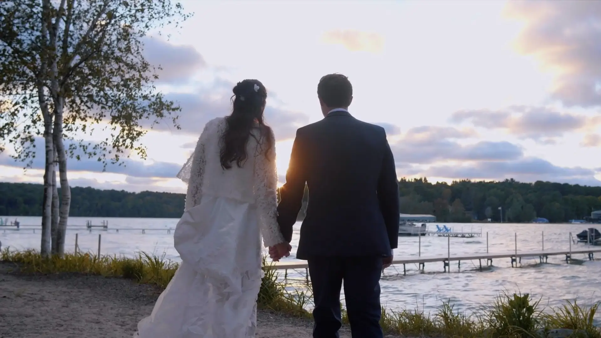 Chloe and Joe hold hands by a beautiful Michigan waterfront during sunset golden hour, captured in this timeless moment by their wedding videographer.