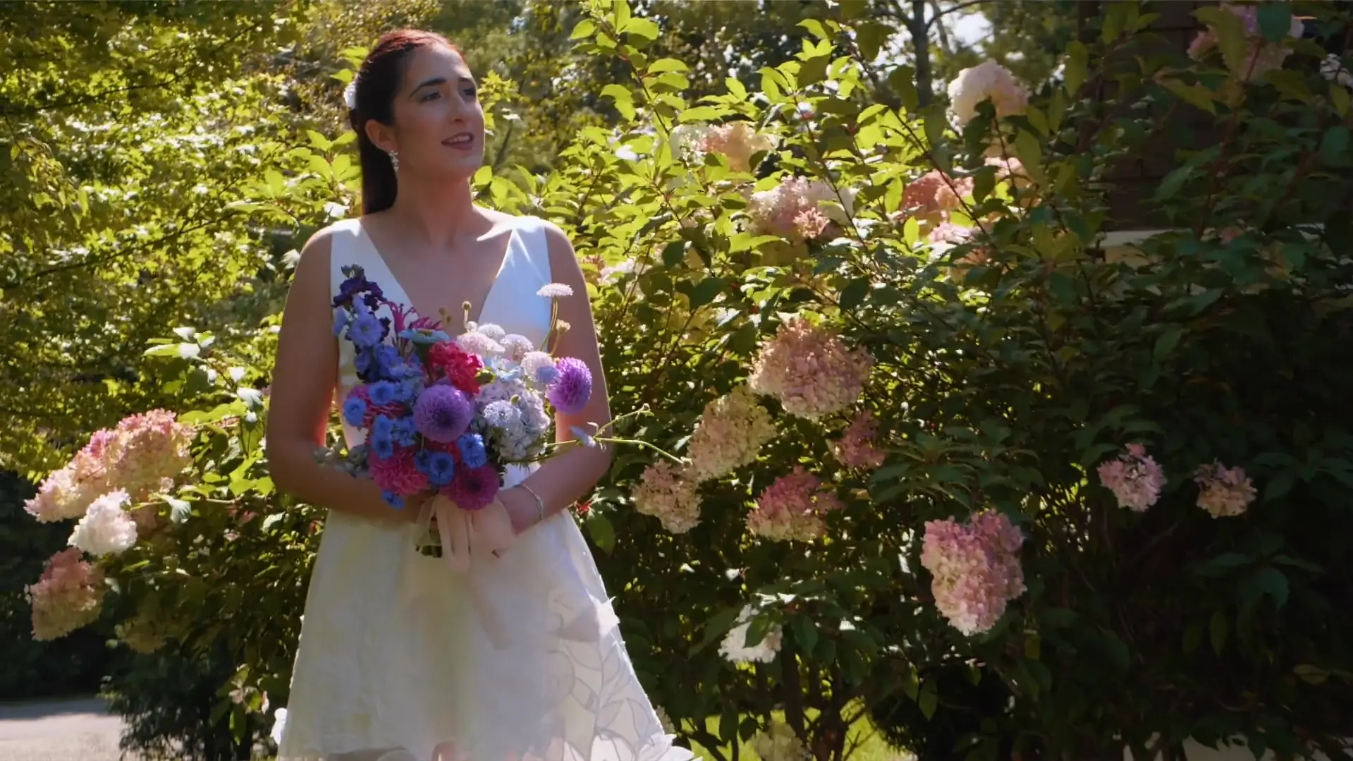 Bride Chloe stands outdoors holding a vibrant bouquet of pink, purple, and blue flowers. She is smiling, surrounded by lush greenery and pink hydrangeas, on a sunny day at Walloon Lake.