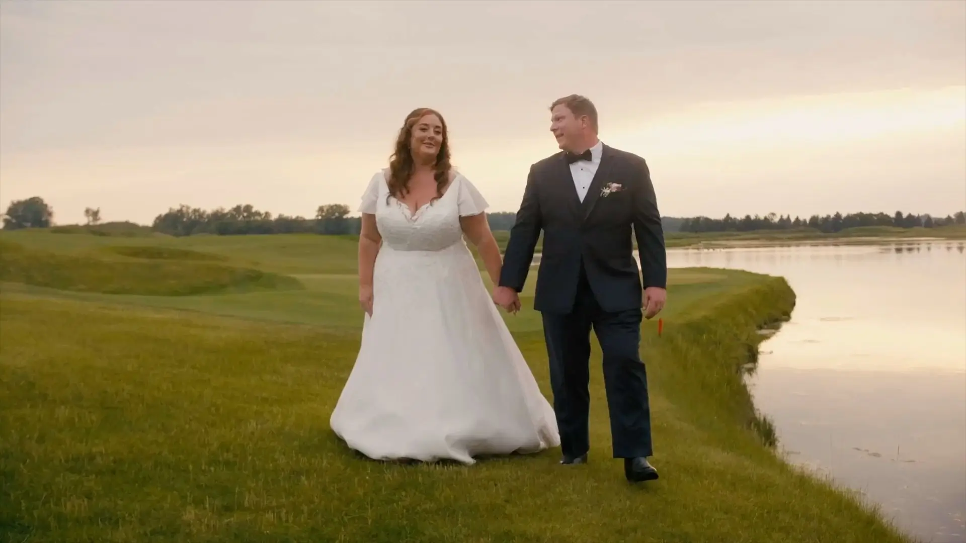 Amanda and Zach walking hand-in-hand on a grassy area near a serene body of water at sunset, with gentle smiles and gazes towards each other. The backdrop includes open fields and the soft glow of the evening sky, adding a romantic ambiance to the moment.
