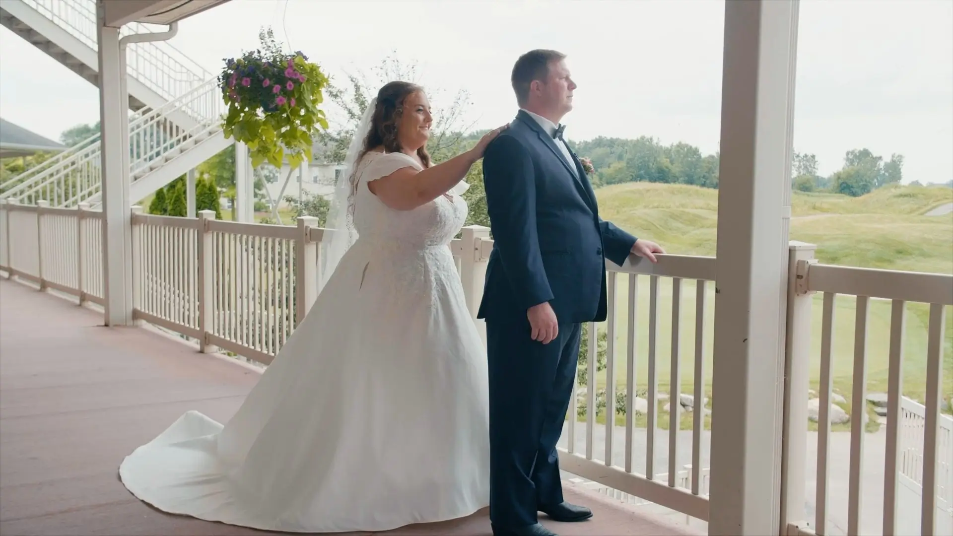 The bride, Amanda, gently touches the groom, Zach's, shoulder from behind during their first look at Eagle Eye Golf & Banquet Center in Bath Township, Michigan. The groom faces away, and they stand on a balcony overlooking the golf course.