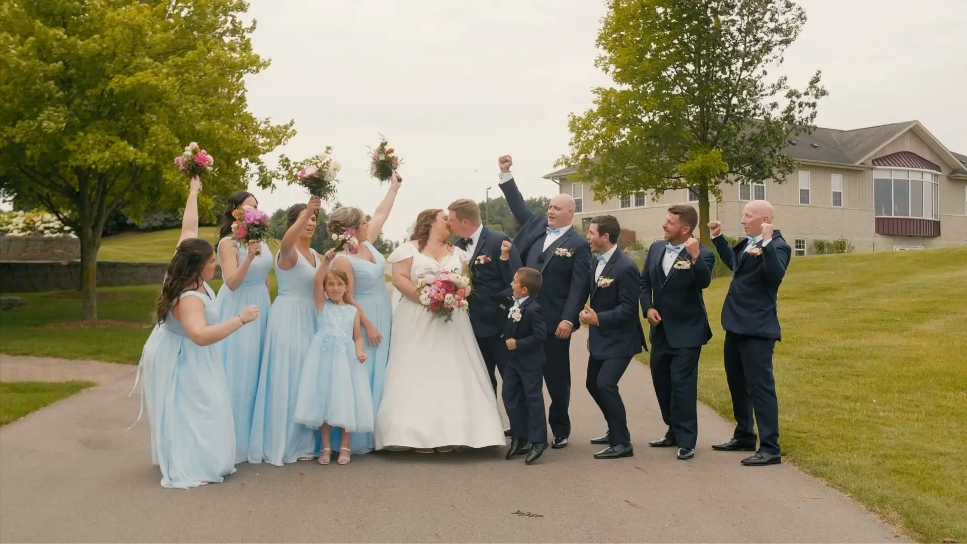 The wedding party of Amanda and Zach celebrates as the couple kisses at Eagle Eye Golf & Banquet Center in Bath Township. Bridesmaids in light blue dresses and groomsmen in dark suits cheer, with the flower girl and ring bearer in the foreground.