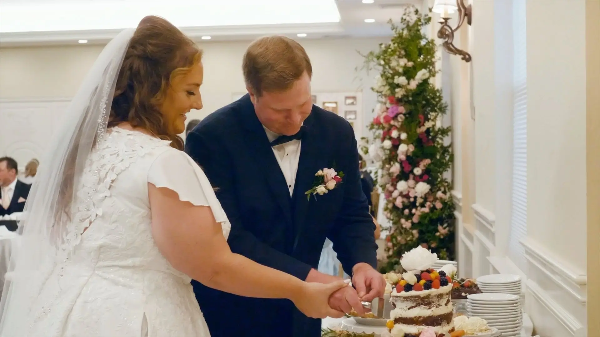 Bride Amanda and groom Zach are cutting their wedding cake together at their reception at Eagle Eye Golf & Banquet Center in Bath Township, Michigan. Both are smiling and wearing traditional wedding attire. The background features decorative flowers.