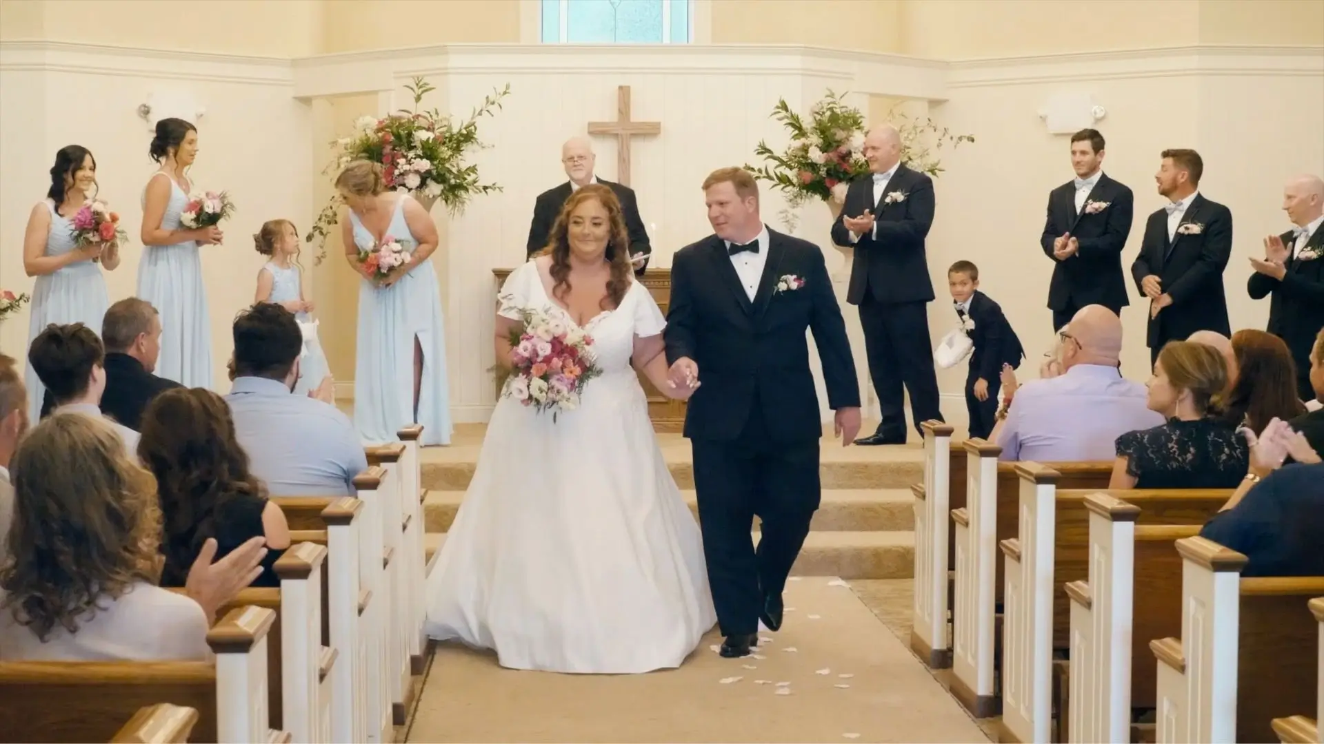 Amanda and Zach walk down the aisle after their wedding ceremony at Hawk Hollow Chapel, Bath Township, Michigan. The bride holds a bouquet of pink and white flowers, accompanied by bridesmaids in light blue dresses and groomsmen in black suits.
