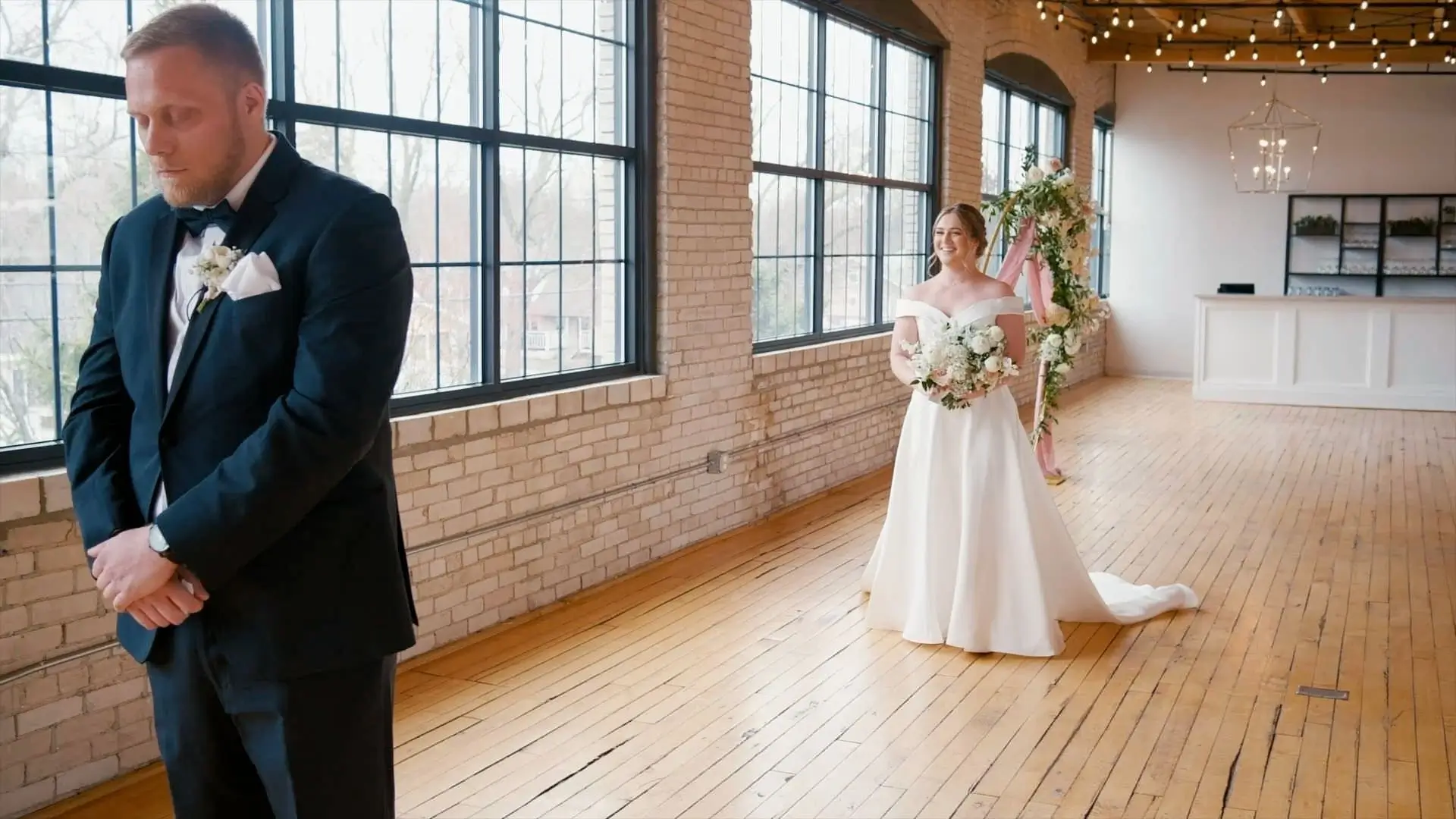 Bride Anna, in an off-the-shoulder white wedding dress holding a bouquet, approaches groom Kevin, in a dark suit with a boutonniere, standing with his eyes closed in a loft with large windows and string lights at a venue in Holland.