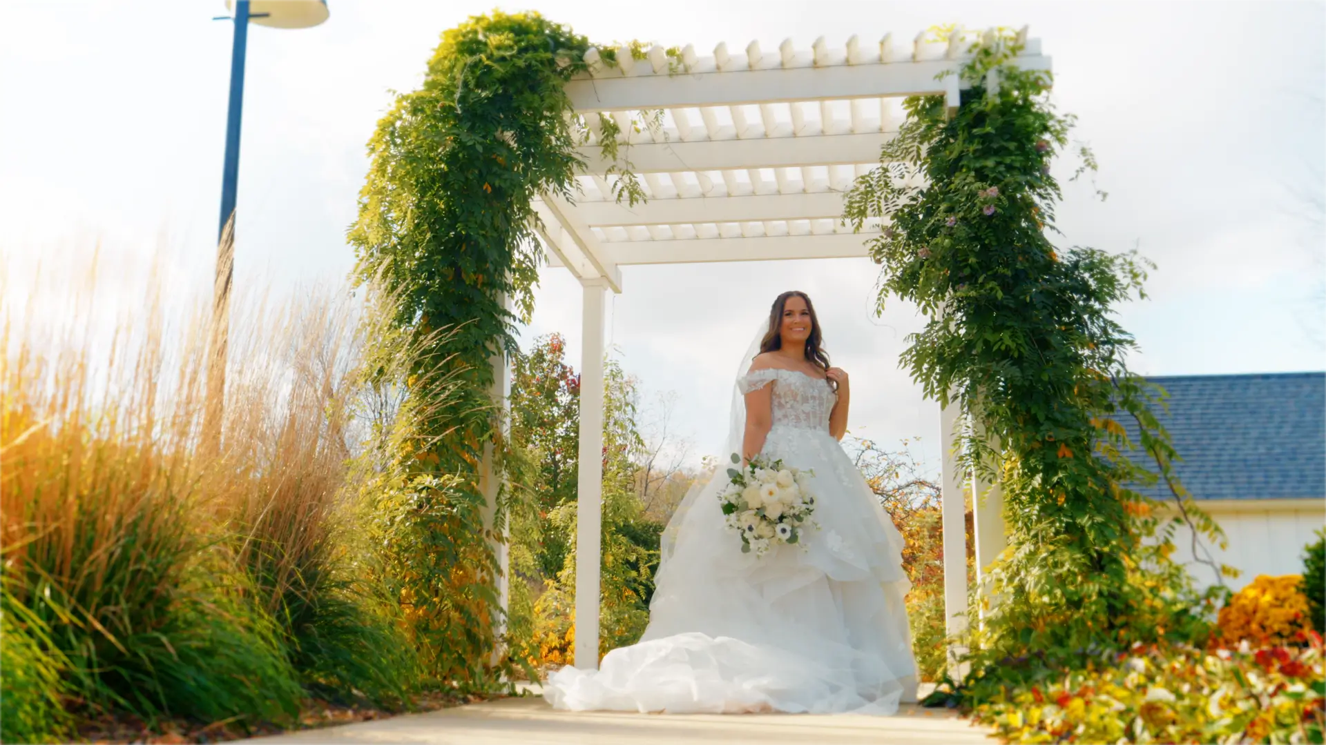 Bride in a white gown stands under a pergola adorned with greenery, holding a bouquet of white flowers. The background features tall grasses and a clear sky, capturing a serene, sunny day at Bay Pointe Inn, Shelbyville, Michigan.