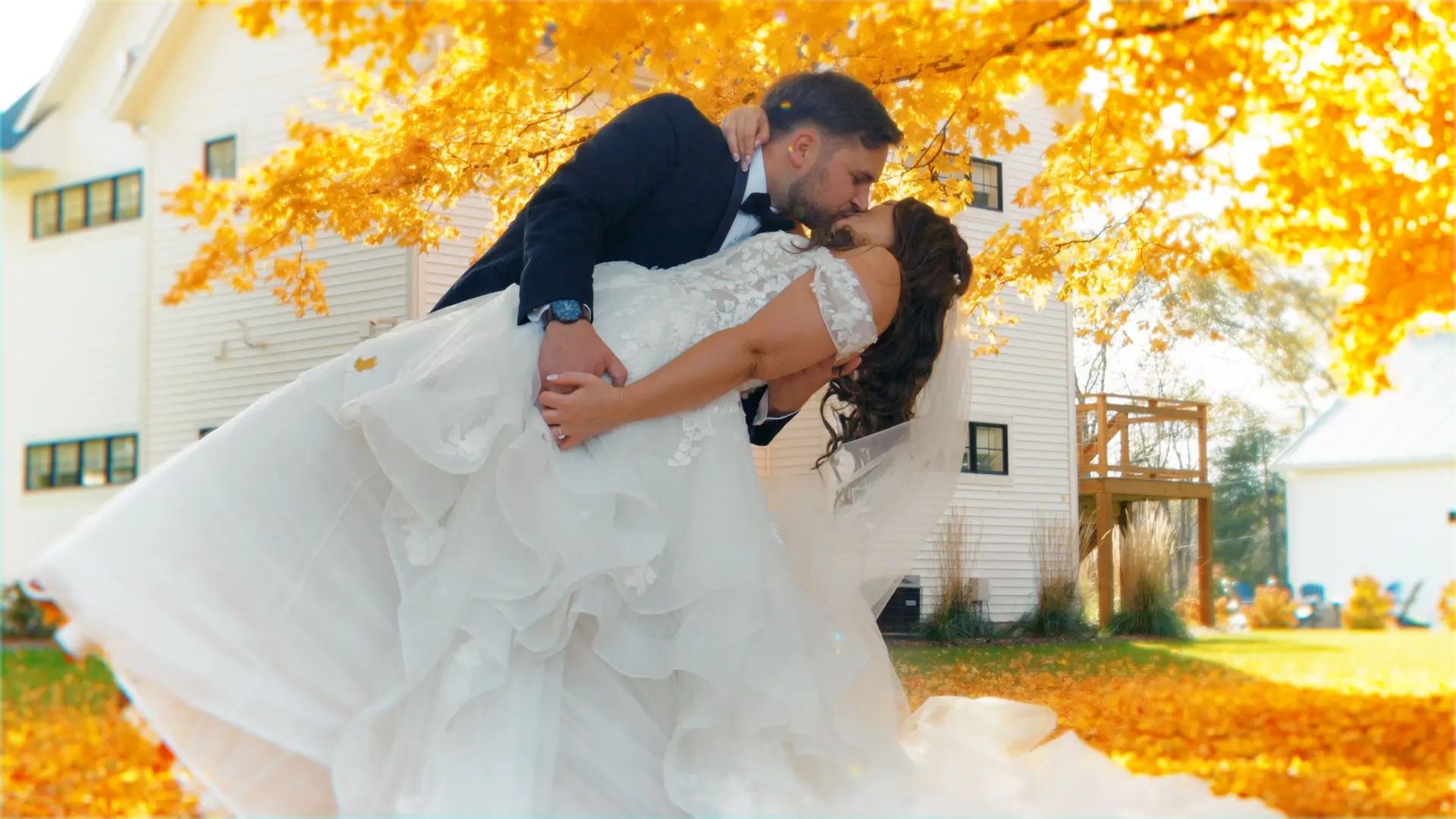 The groom dips the bride for a kiss under vibrant autumn leaves, with a white building and golden foliage in the background at Bay Pointe Inn, Shelbyville, Michigan.
