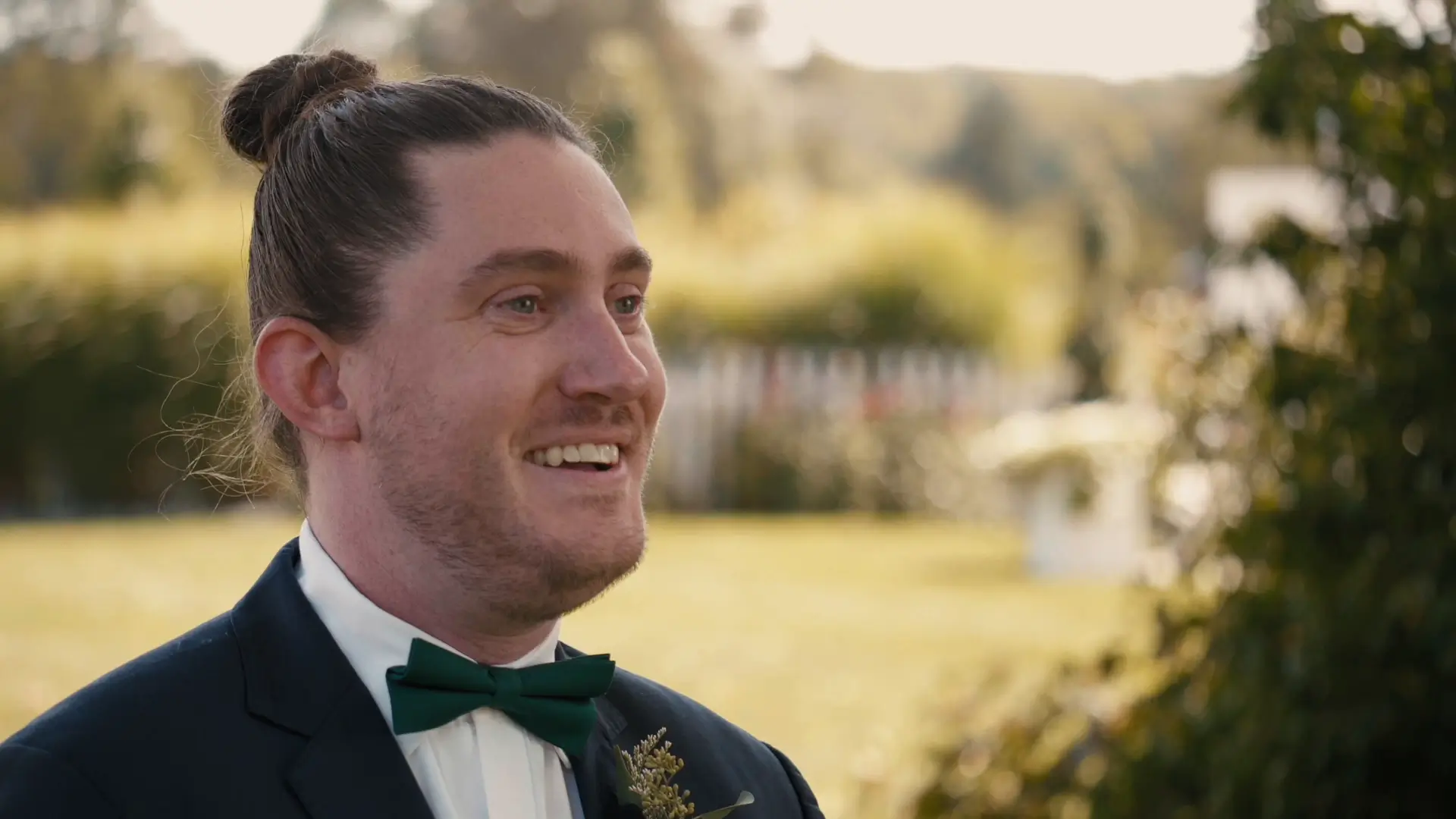 A groom in a suit with a green bow tie smiles excitedly during his first look outdoors against a blurred sunny garden background. His wedding videographer captured the moment he first saw his bride in her dress before their wedding ceremony.
