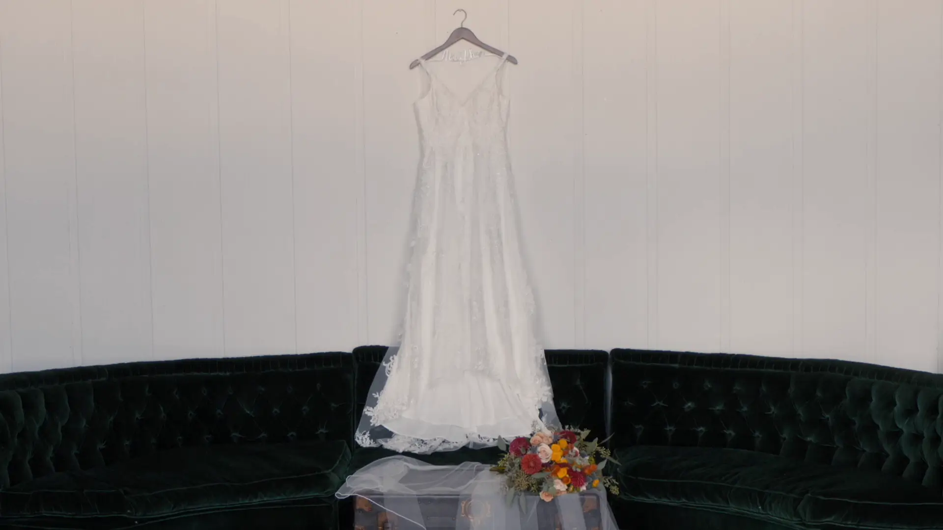 Taylor's white wedding dress hangs on a dark hanger against a light wall, above a green velvet couch. A table beneath holds a veil and a colorful bouquet with red, pink, and yellow flowers.
