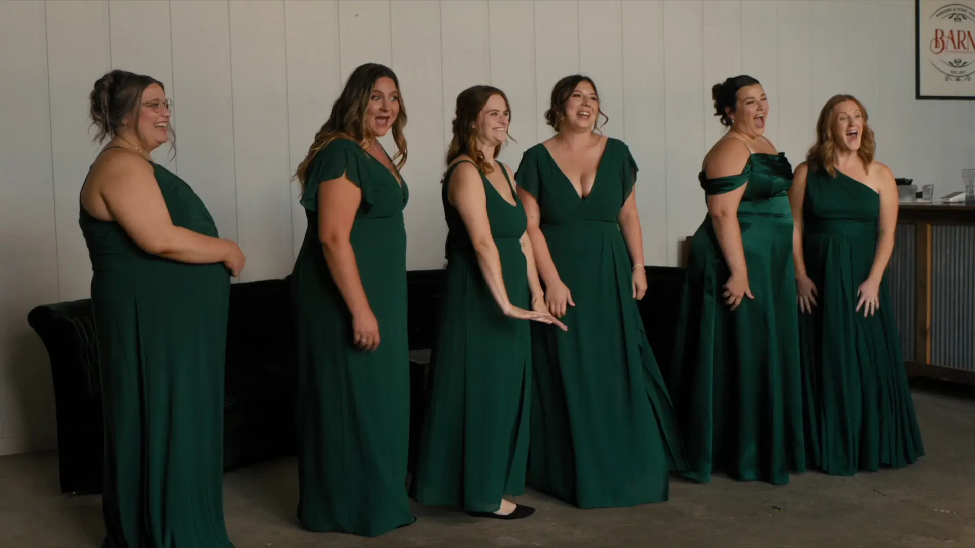 Bridesmaids in dark green dresses stand indoors with expressions of surprise and joy as they get a first look at the bride in her final hair and makeup at Barn 1888 in the Hopkins and Allegan area, Michigan.