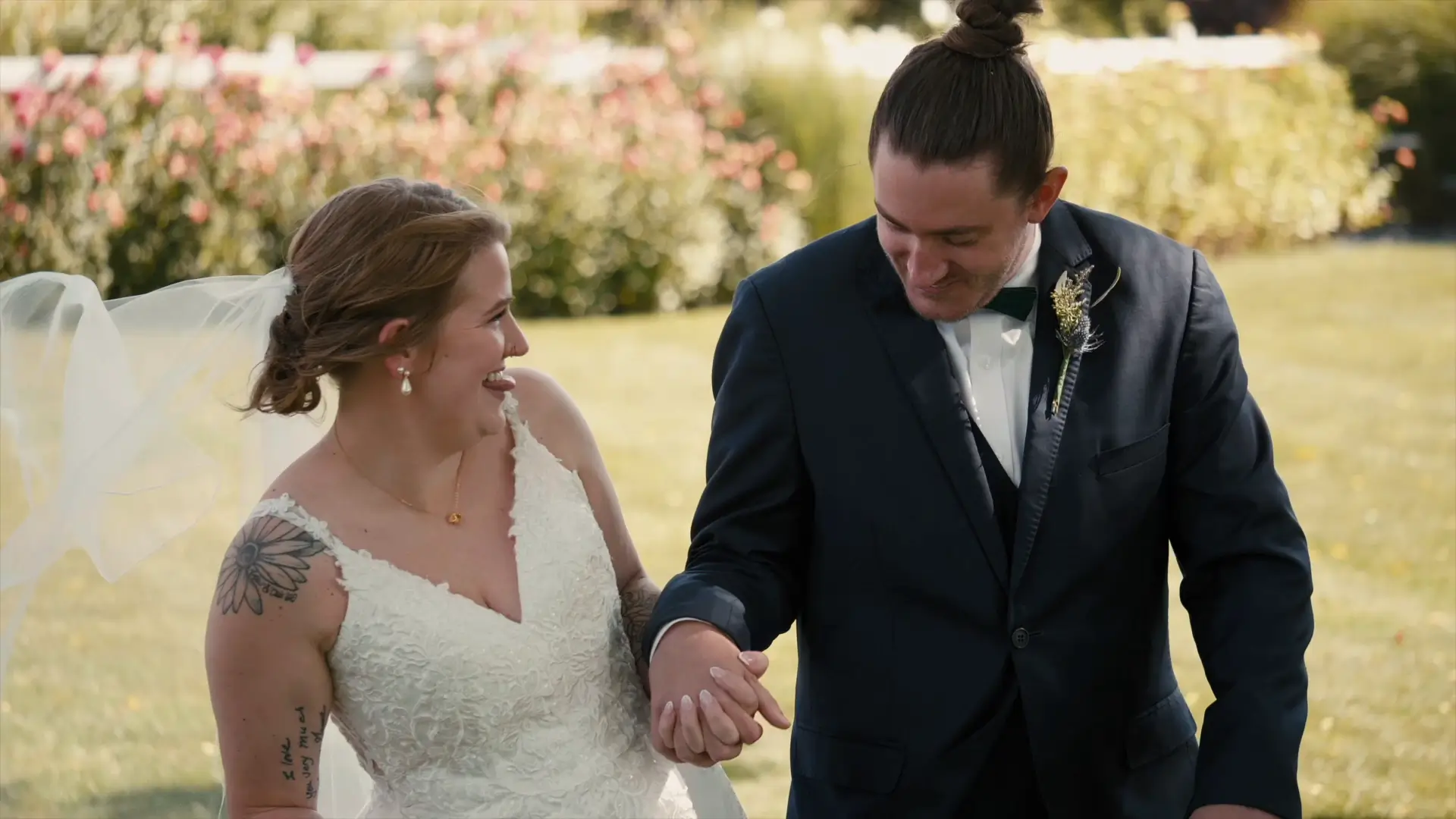 The bride and groom are smiling and holding hands outdoors in a garden setting, with the bride in a white lace dress and veil and the groom in a navy suit with a boutonniere. Their expressions are silly and joyful, as captured for their wedding video.