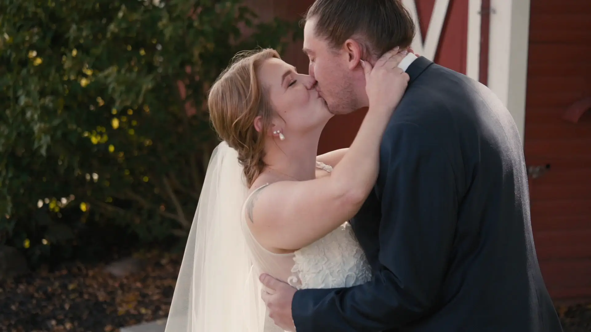 Bride and groom share a kiss outdoors in front of a rustic barn backdrop. The bride wears an elegant veil and lace gown, while the groom is in a dark suit. Nearby greenery adds a touch of natural beauty, enhancing the romantic atmosphere.