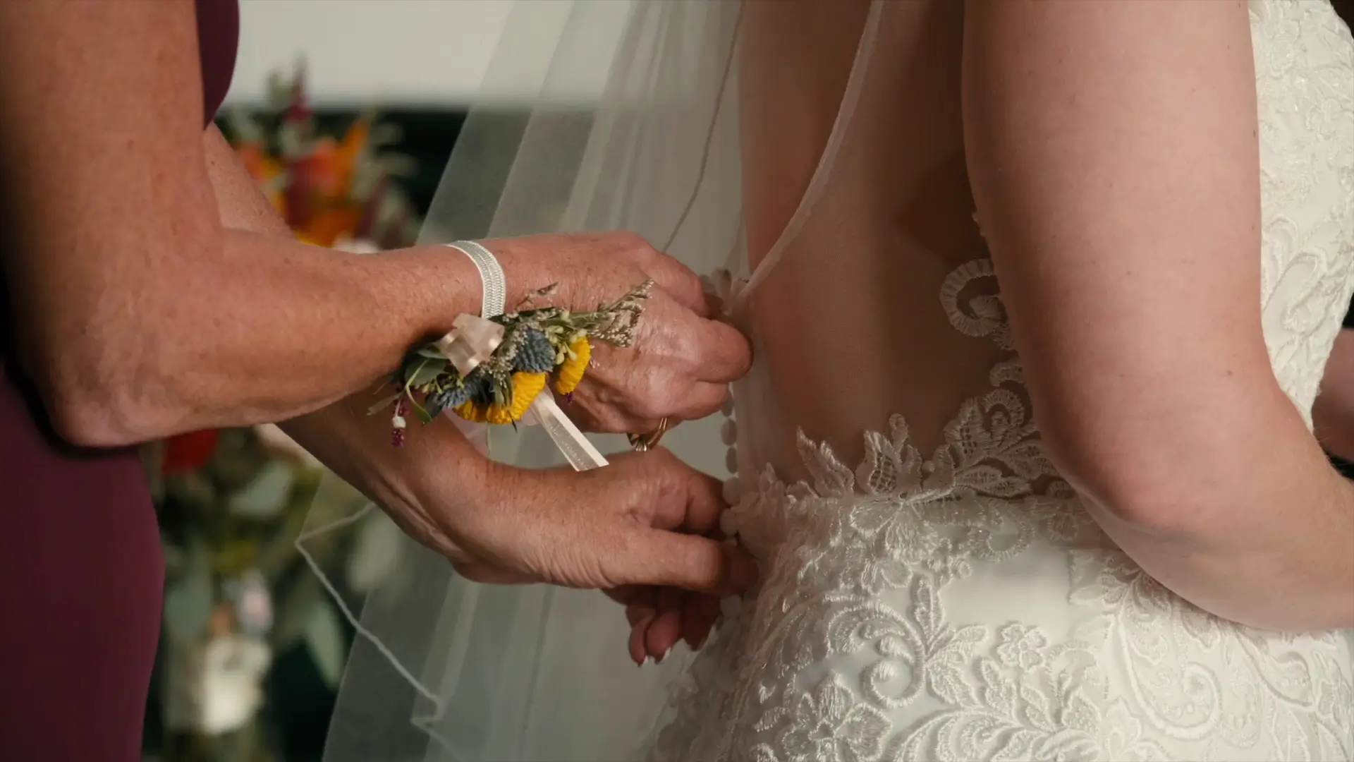 This close-up moment shows the bride's mother fastening buttons on the back of the bride's lace wedding gown. The bride's veil is visible, and the woman wears a floral wrist corsage.